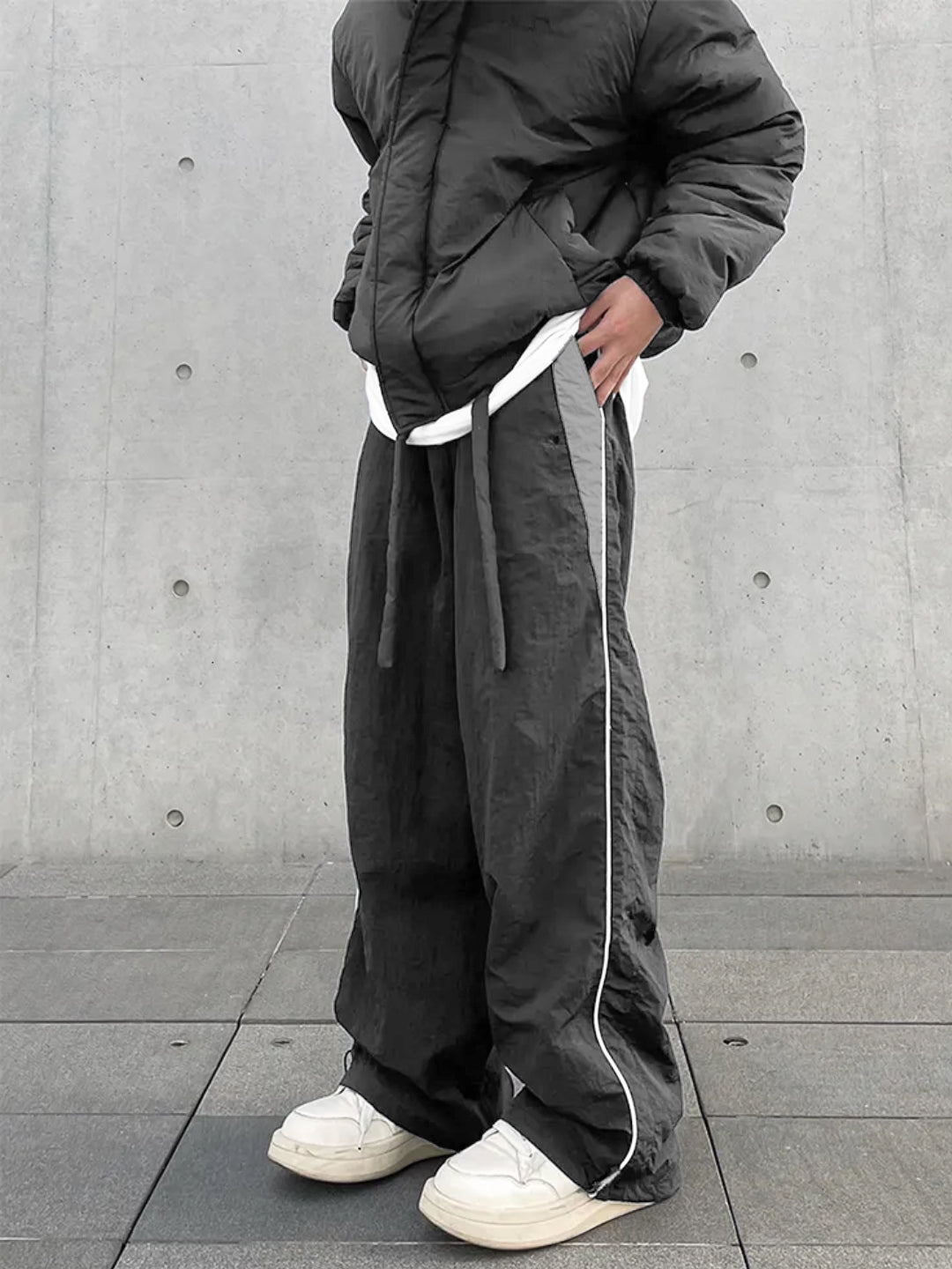 A person wearing dark grey trackpants with contrast white piping and an elasticated waistband with drawstrings. The person is standing against a concrete background wearing white sneakers.