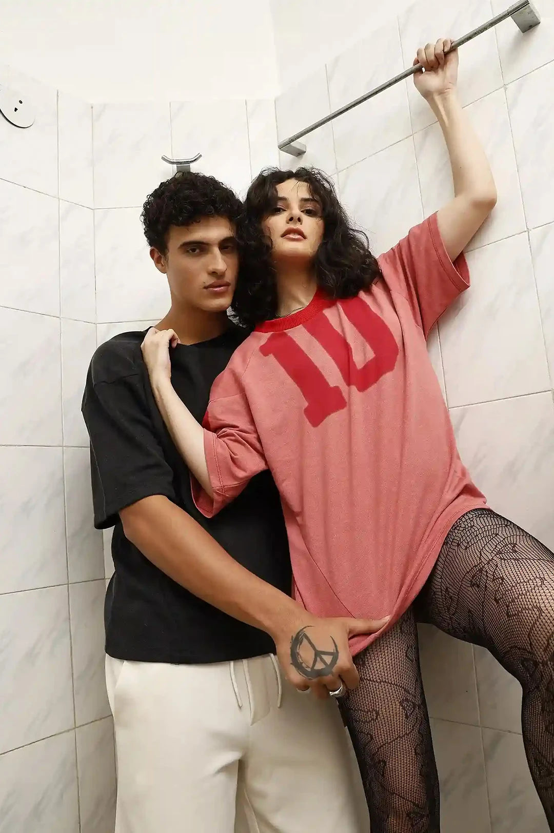 Two people standing in a tiled room, one wearing a black shirt and white pants, the other in a red 'UP' shirt and fishnet stockings.
