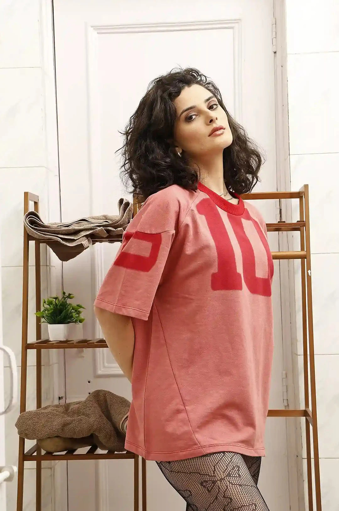 Woman wearing a pink t-shirt with red text in a room with wooden shelves and plants.