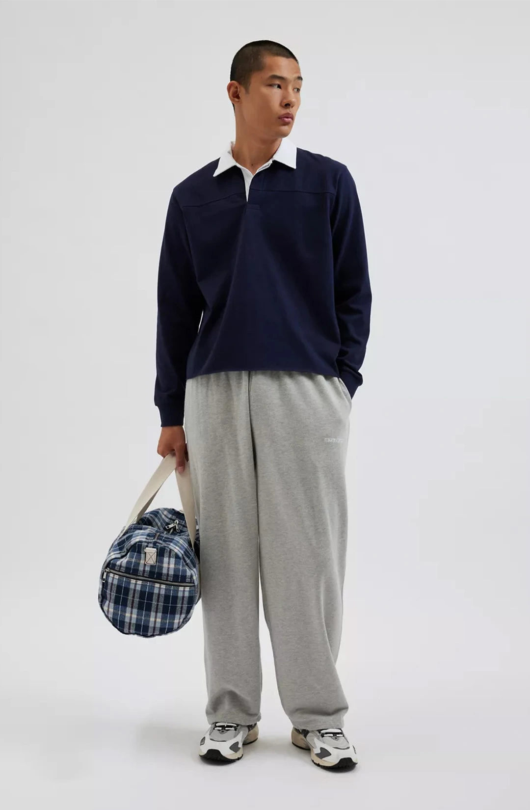 Man holding a checkered bag wearing a navy sweater and gray pants on a white background
