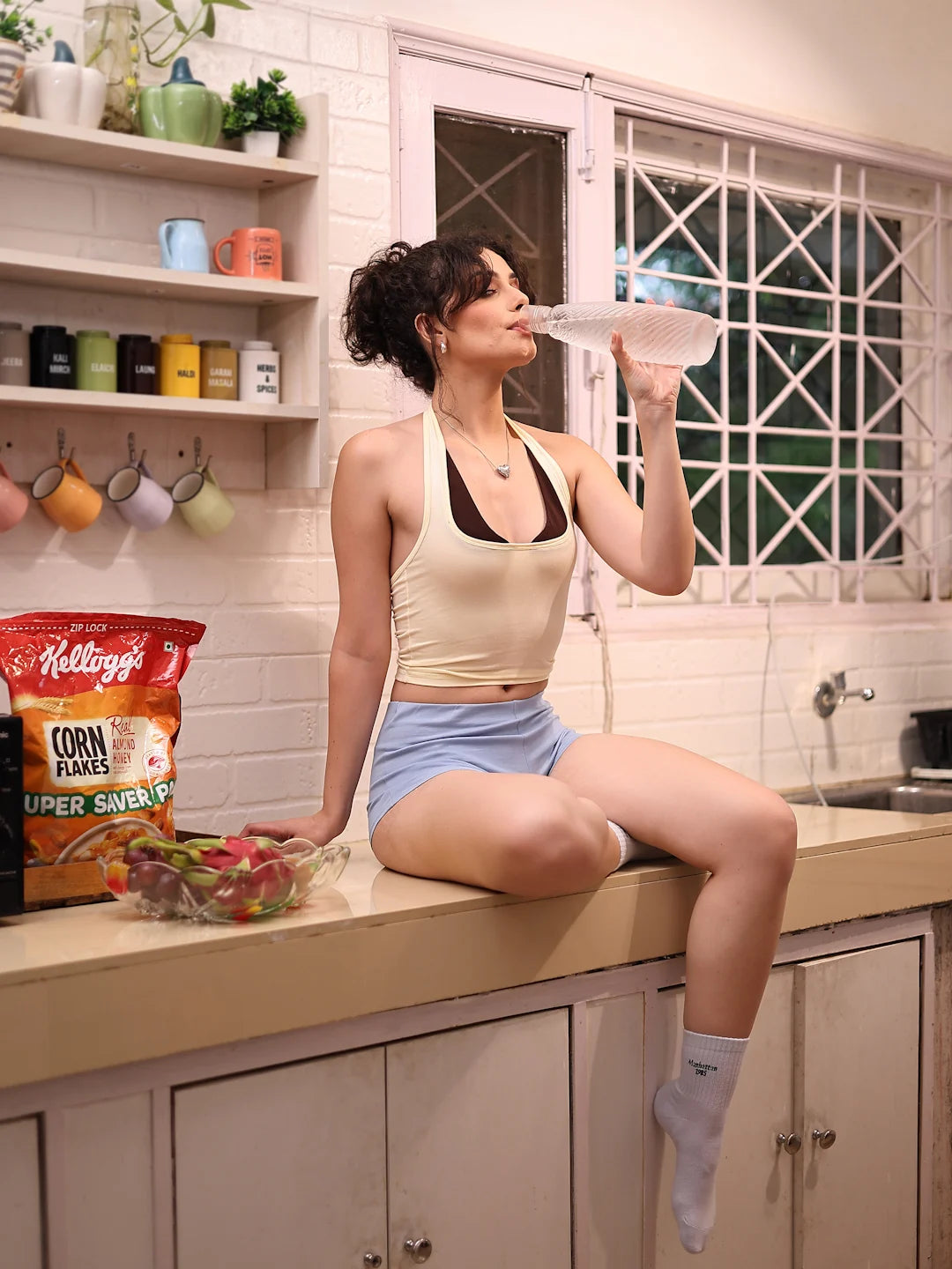 Woman sitting on a kitchen counter holding a bowl, with Kellogg's Corn Flakes and fruit on the counter.