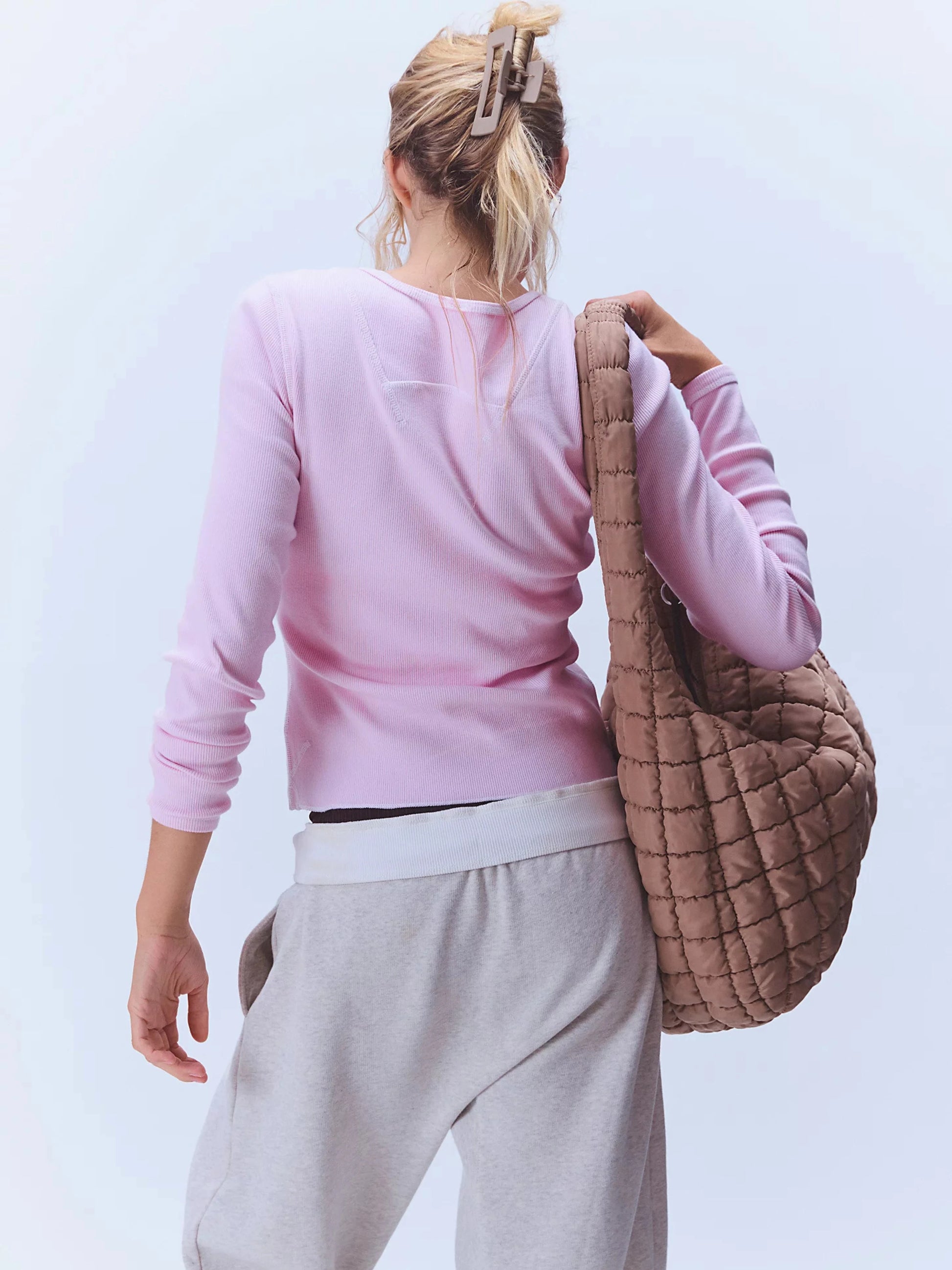 Person wearing a pink long-sleeve top and gray pants, holding a brown quilted bag against a light background
