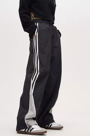 A person standing sideways, wearing black striped parachute pants with white contrast piping, an elasticated waistband with adjustable drawstrings, and white sneakers.