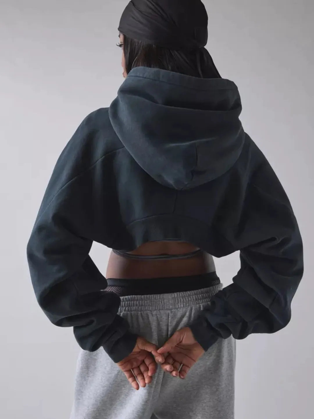 Person wearing a black cropped hoodie with cut-out design over a sports bra and shorts on a plain background