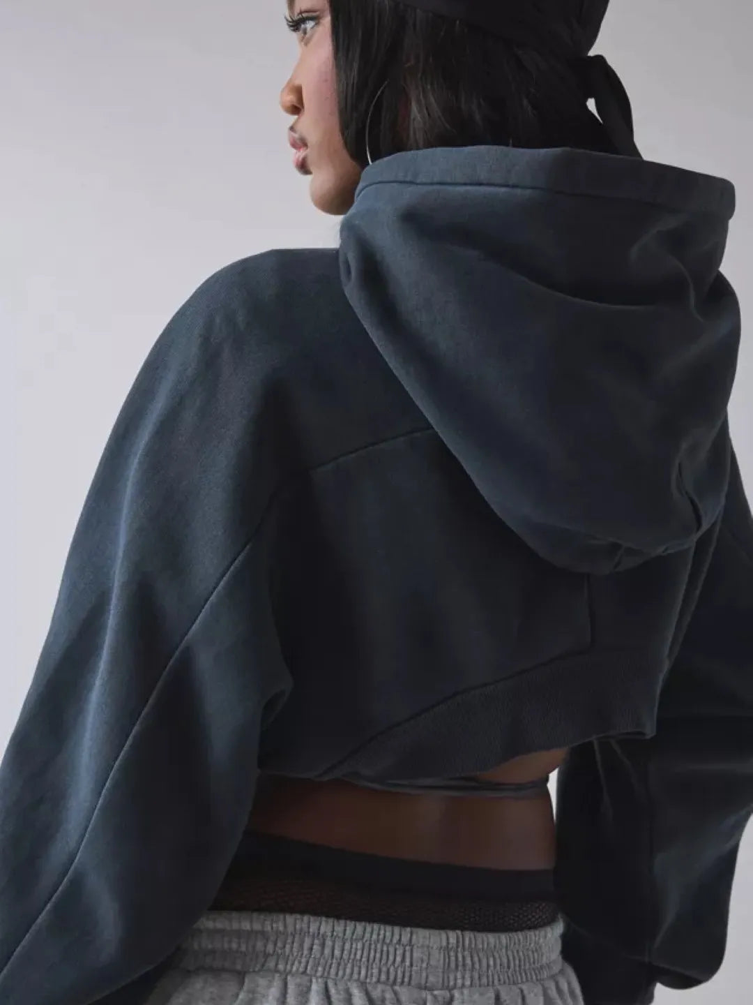 Person wearing a black cropped hoodie with cut-out design over a sports bra and shorts on a plain background