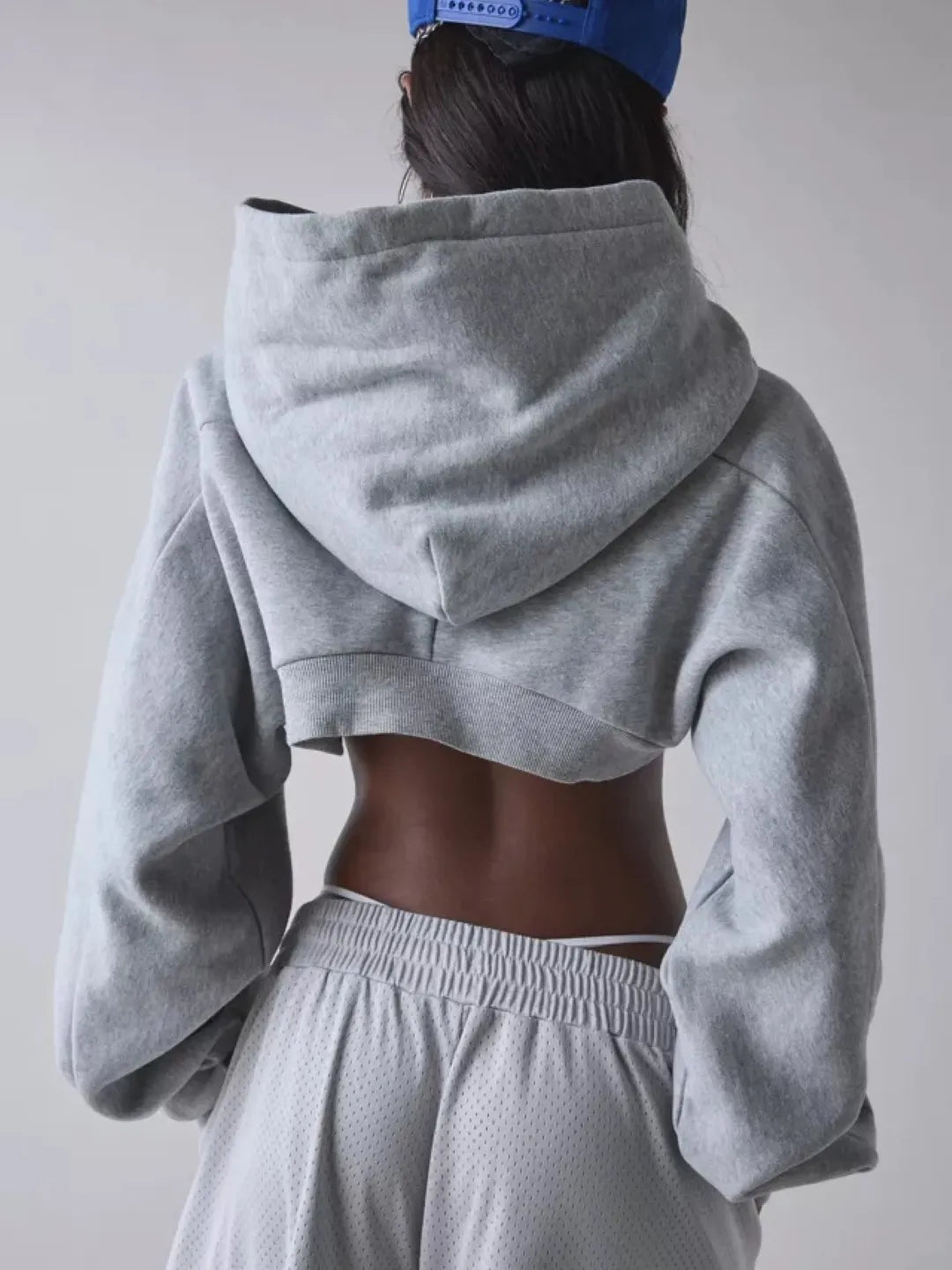 Person wearing a gray cropped hoodie with cut-out design over a sports bra and shorts on a plain background