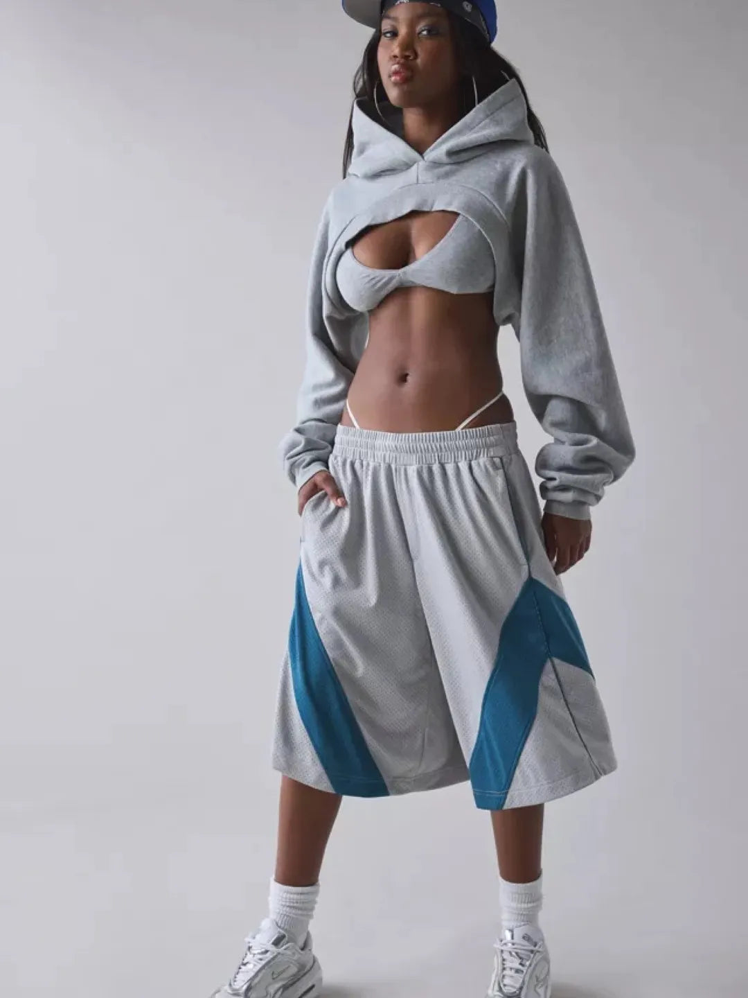 Person wearing a gray cropped hoodie with cut-out design over a sports bra and shorts on a plain background