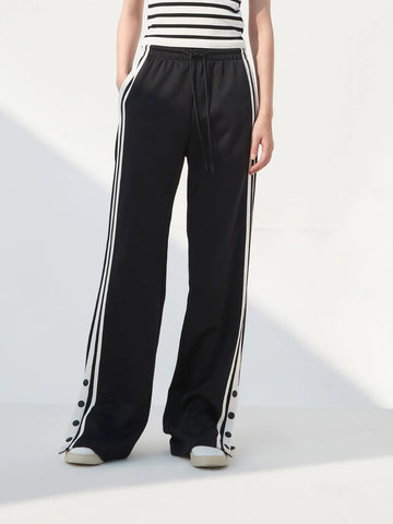A pair of black slit snap button tracks pants with an elastic drawstring waist, featuring white stripes on the legs and a side slit on one leg. The model is wearing a white top and beige shoes.