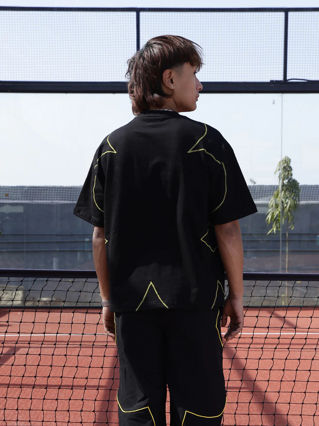 Person wearing a black t-shirt with yellow geometric designs on a tennis court.