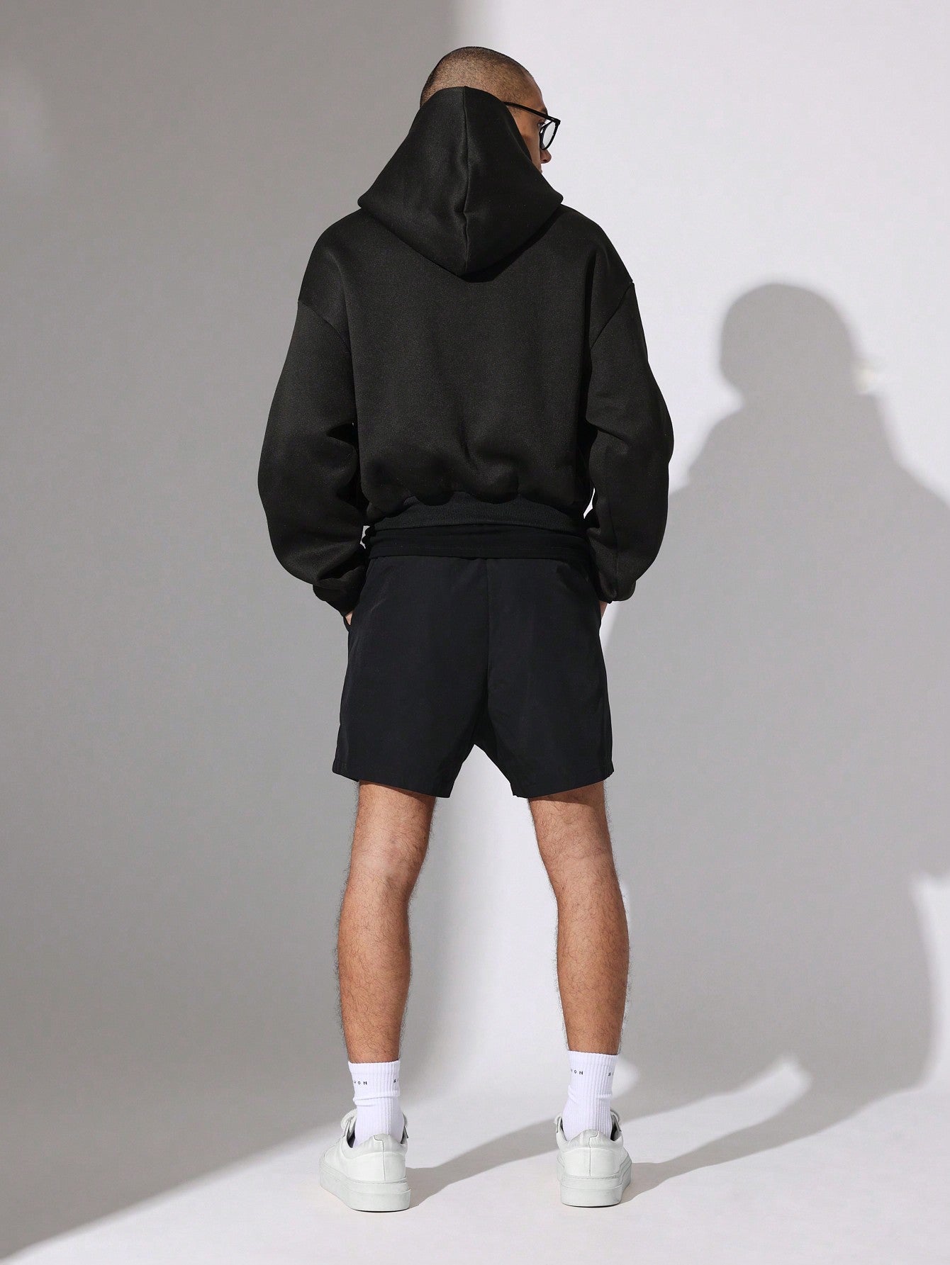 Person wearing a black hoodie and shorts with white sneakers against a plain background
