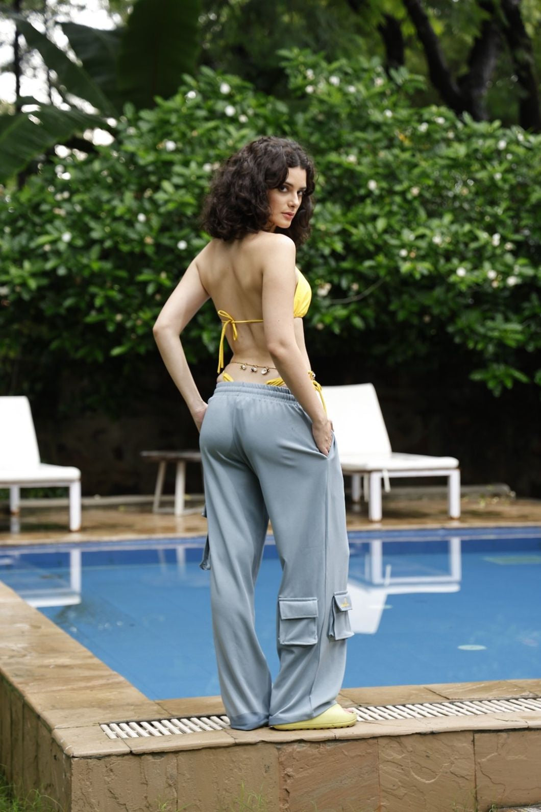Woman in a yellow bikini top and gray pants standing by a pool with greenery in the background