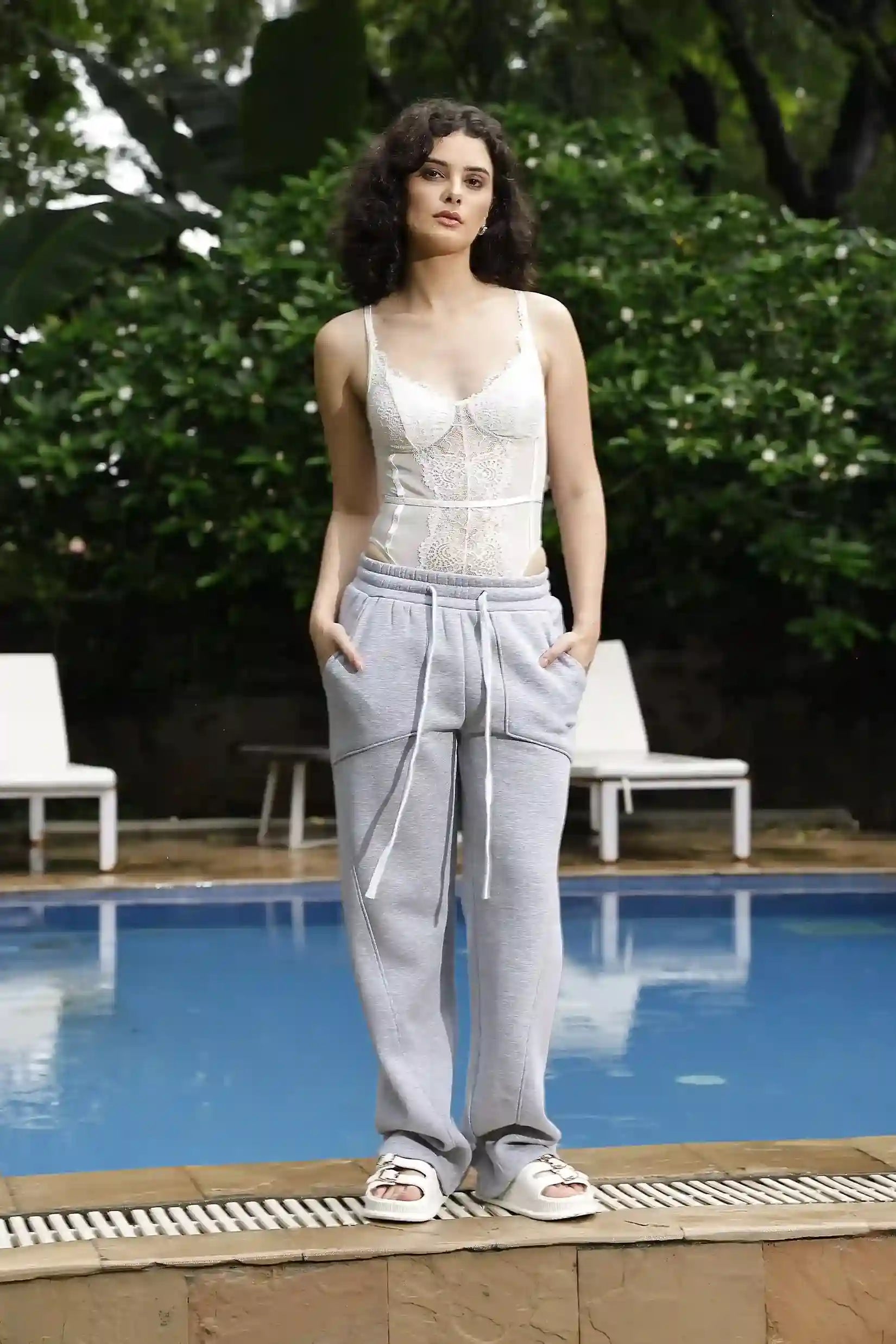 Woman standing by a pool wearing a white lace top and gray pants with greenery in the background.