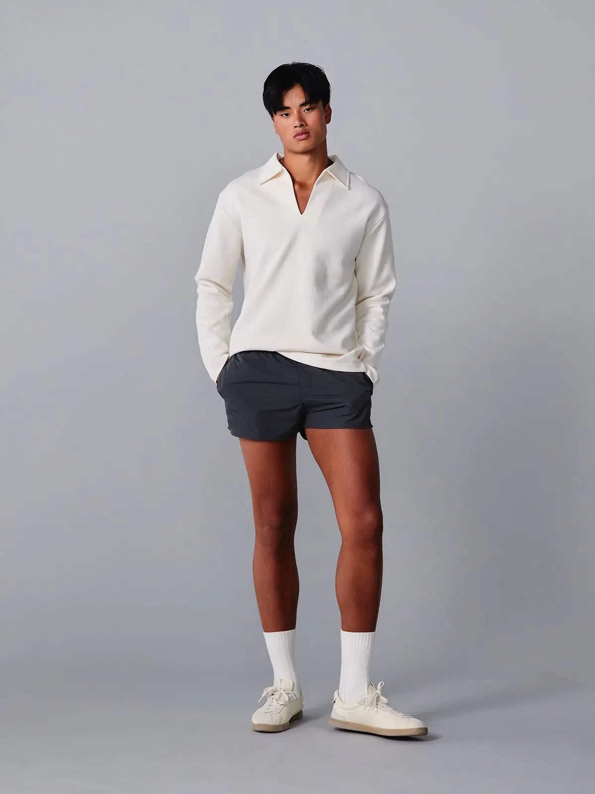Person wearing a white long-sleeve shirt, dark shorts, and white sneakers on a grey background