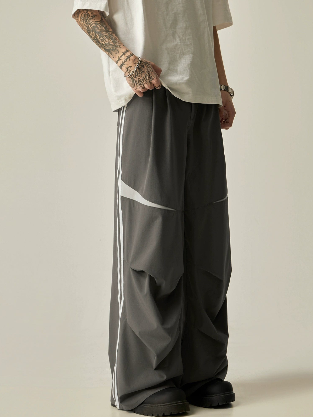 A pair of gray parachute pants with white stripes on the side, worn by a model with a white t-shirt.