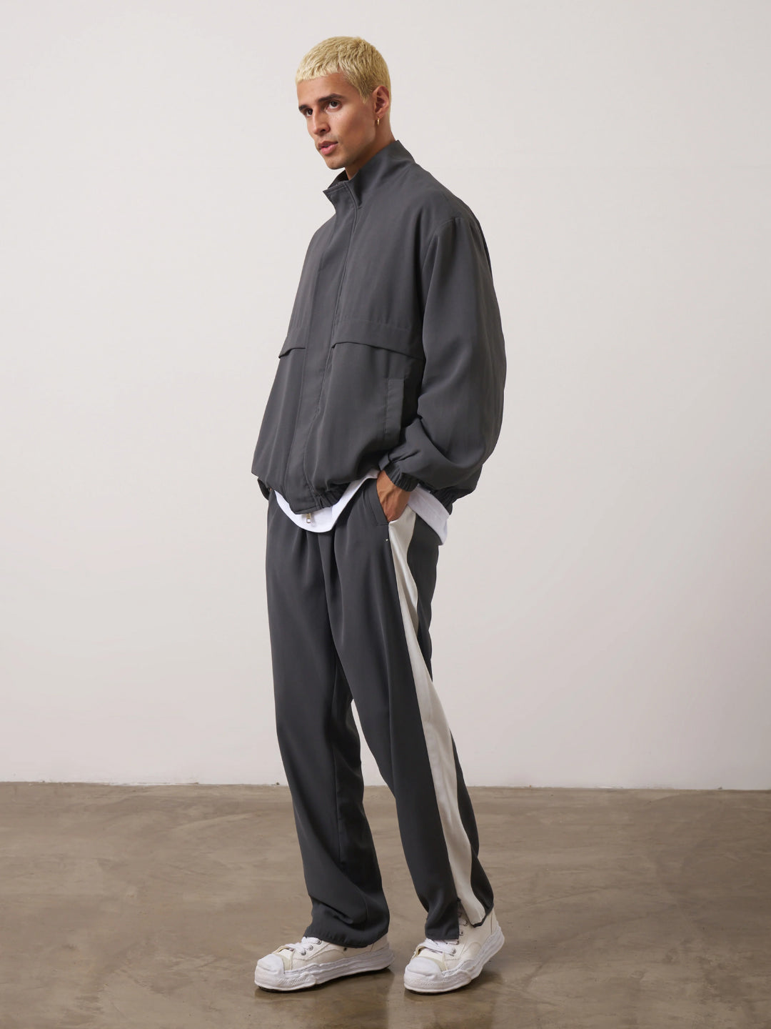 A man standing wearing a dark gray tracksuit, consisting of a jacket with a funnel neck and front pockets, and matching pants with an elasticated waistband.