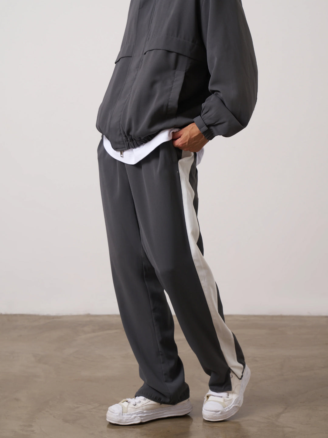 A man standing wearing a dark gray tracksuit, consisting of a jacket with a funnel neck and front pockets, and matching pants with an elasticated waistband.