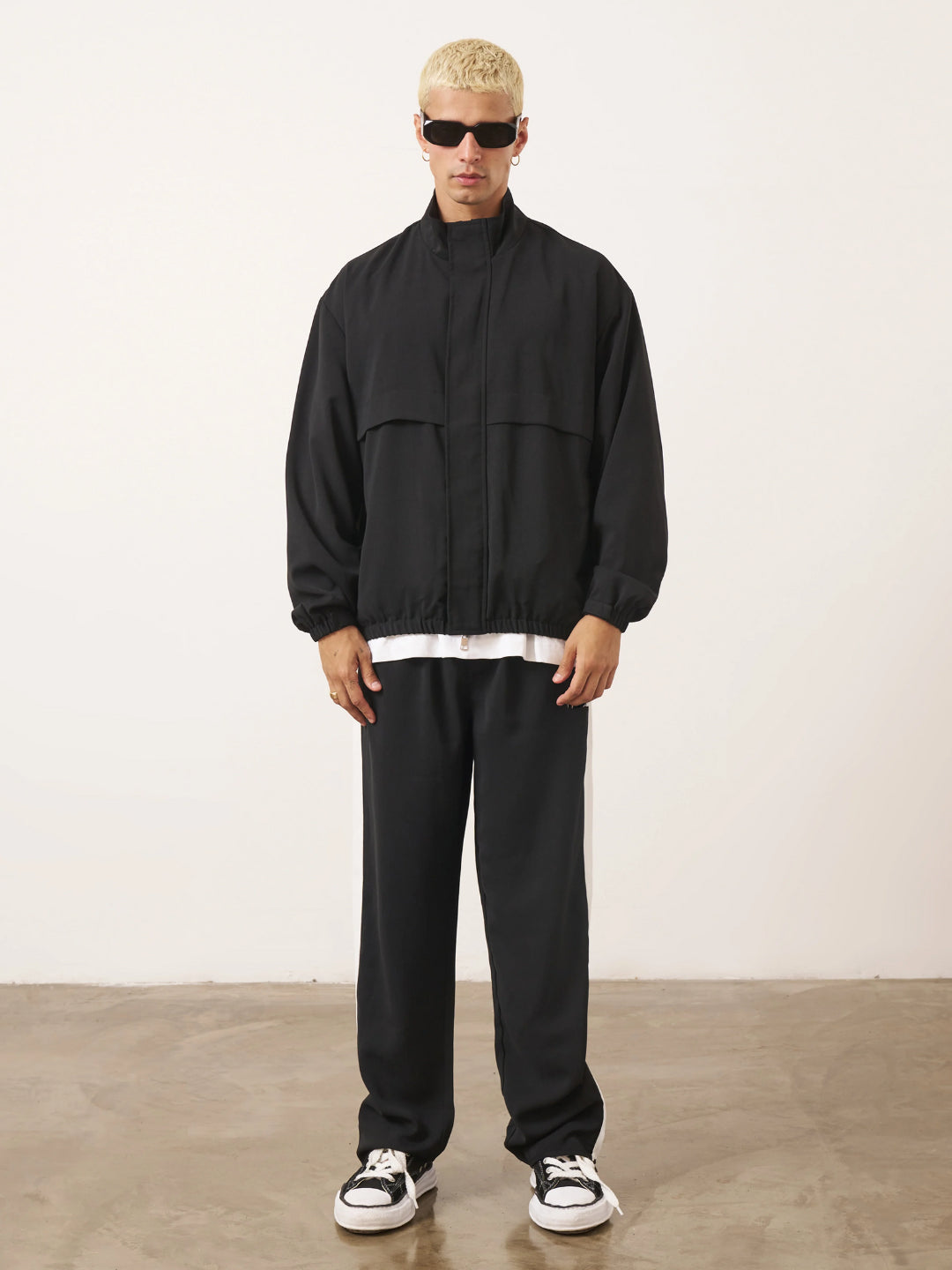 A man standing wearing a black tracksuit, with the jacket featuring a funnel neck and front pockets, and the pants having an elasticated waistband.