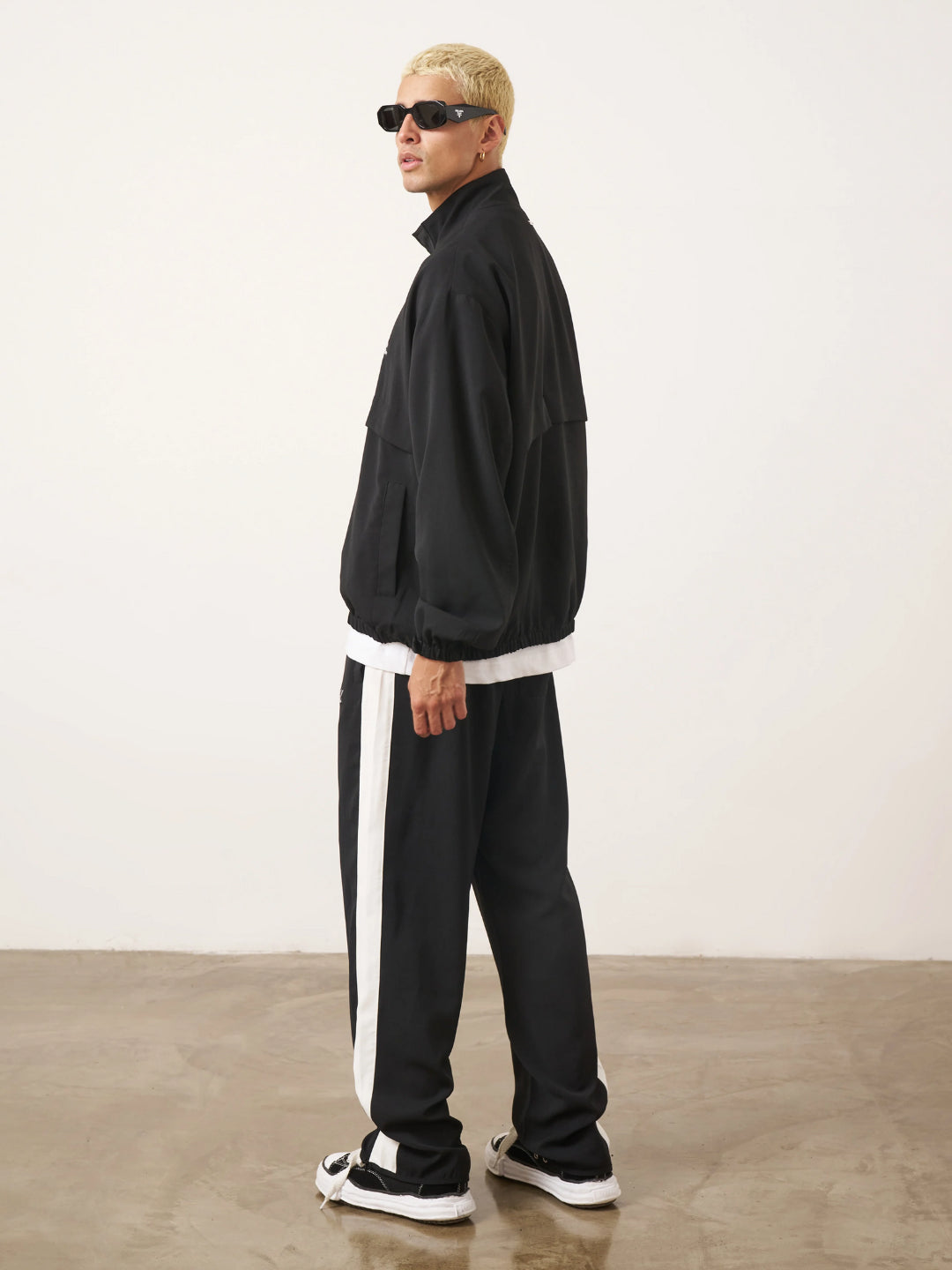 A man standing wearing a black tracksuit, with the jacket featuring a funnel neck and front pockets, and the pants having an elasticated waistband.