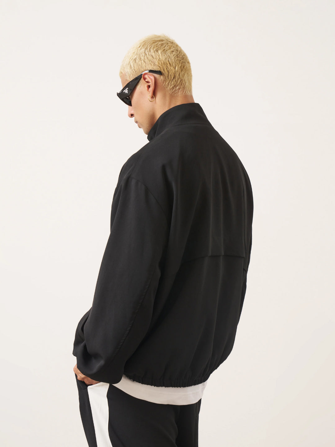 A man standing wearing a black tracksuit, with the jacket featuring a funnel neck and front pockets, and the pants having an elasticated waistband.