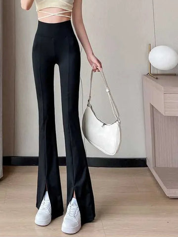 A person wearing high-waist black yoga pants with a front slit, paired with white sneakers, carrying a white bag, with a decorative lamp in the background.