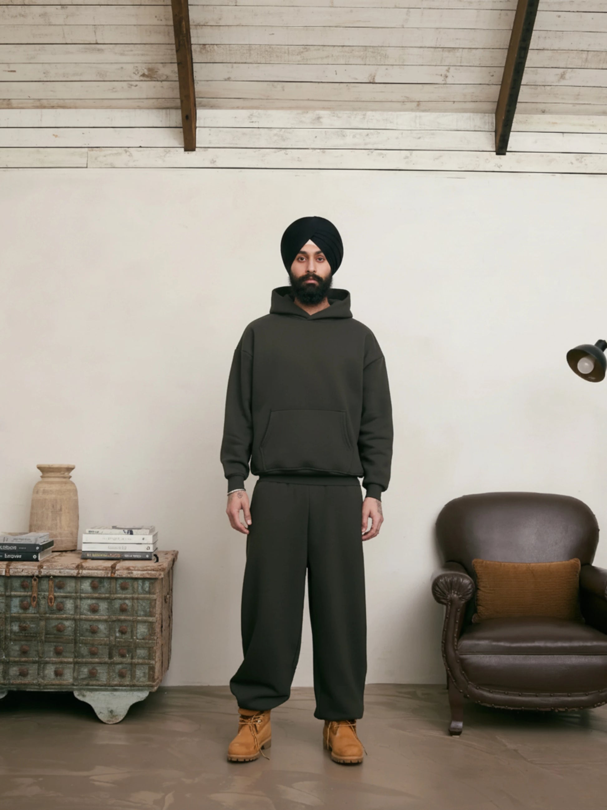 Man wearing a green hoodie and pants standing in a room with a wooden floor and white walls.