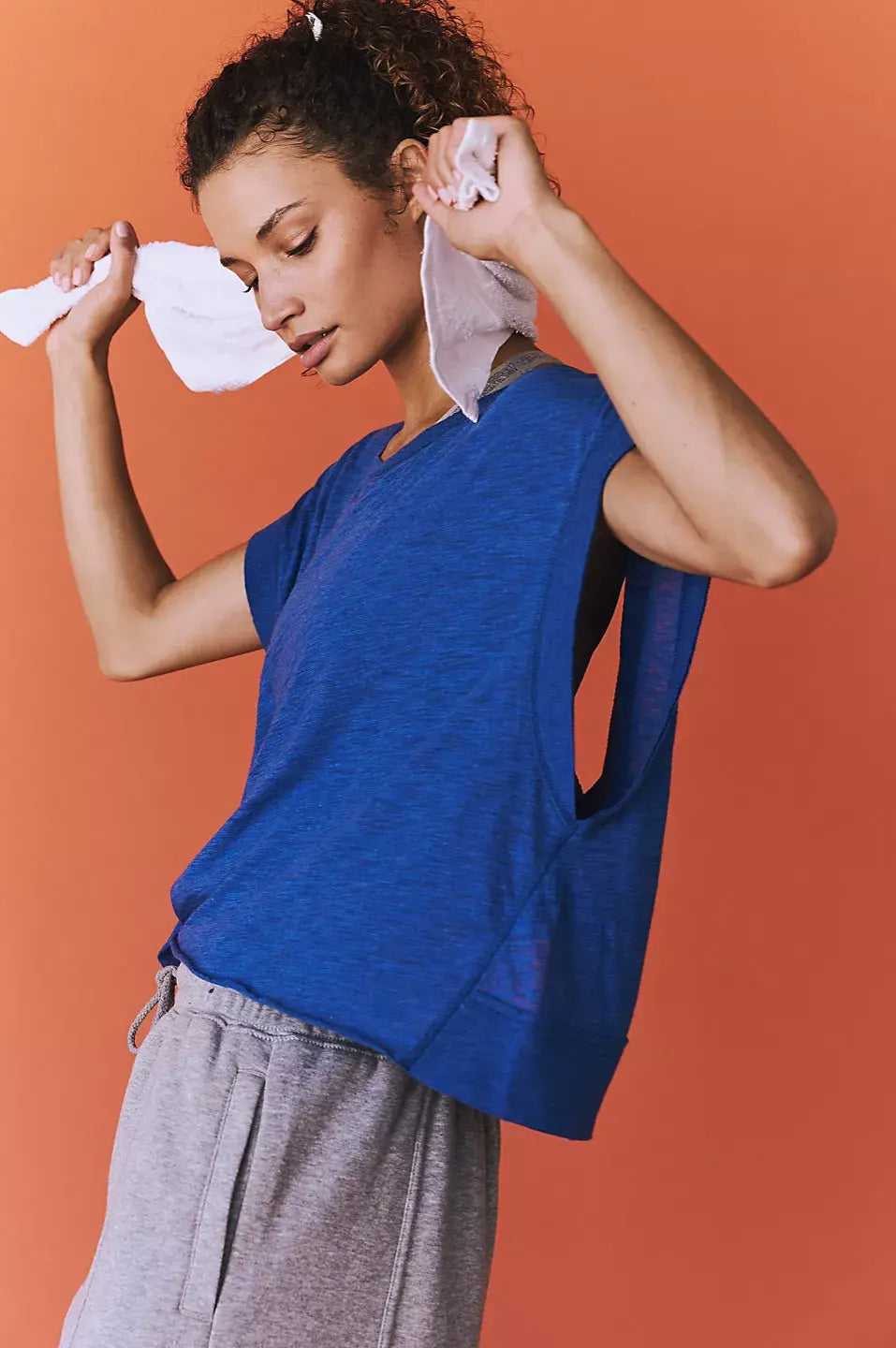 Woman in blue shirt and gray pants wiping face with a towel against an orange background