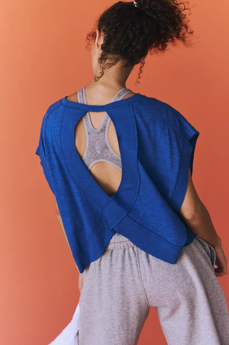 Person wearing a blue top with an open back over a gray sports bra and pants on an orange background