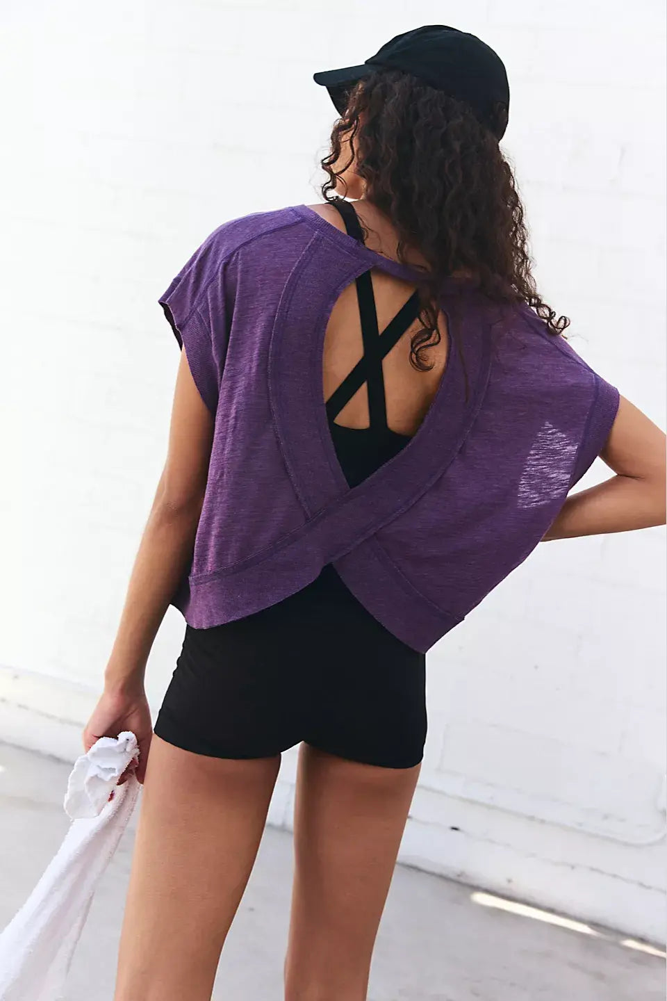 Person wearing a purple top with a black cross design on the back, standing against a white wall.