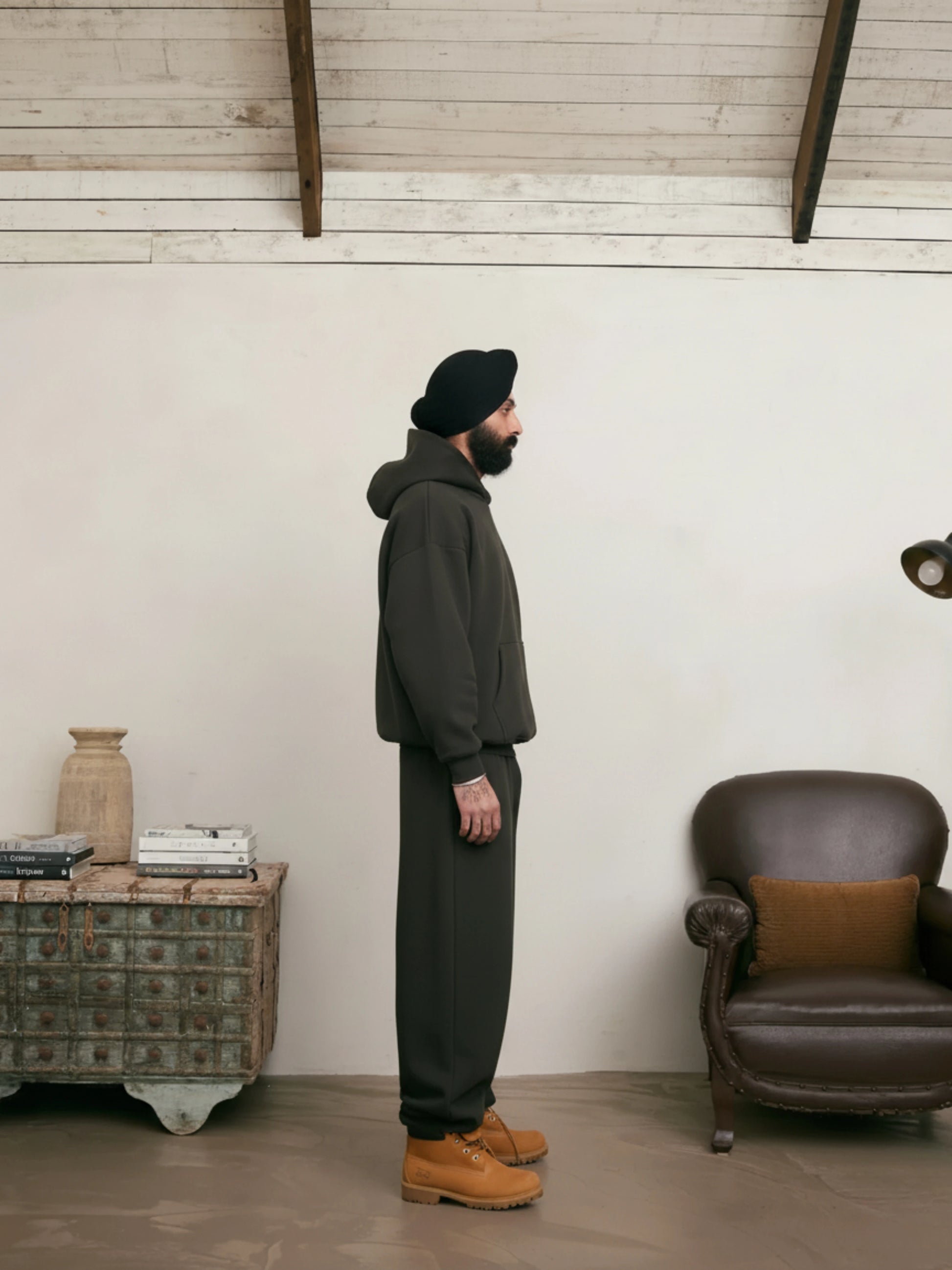 Man in dark outfit standing in a room with a brown armchair and wooden side table.
