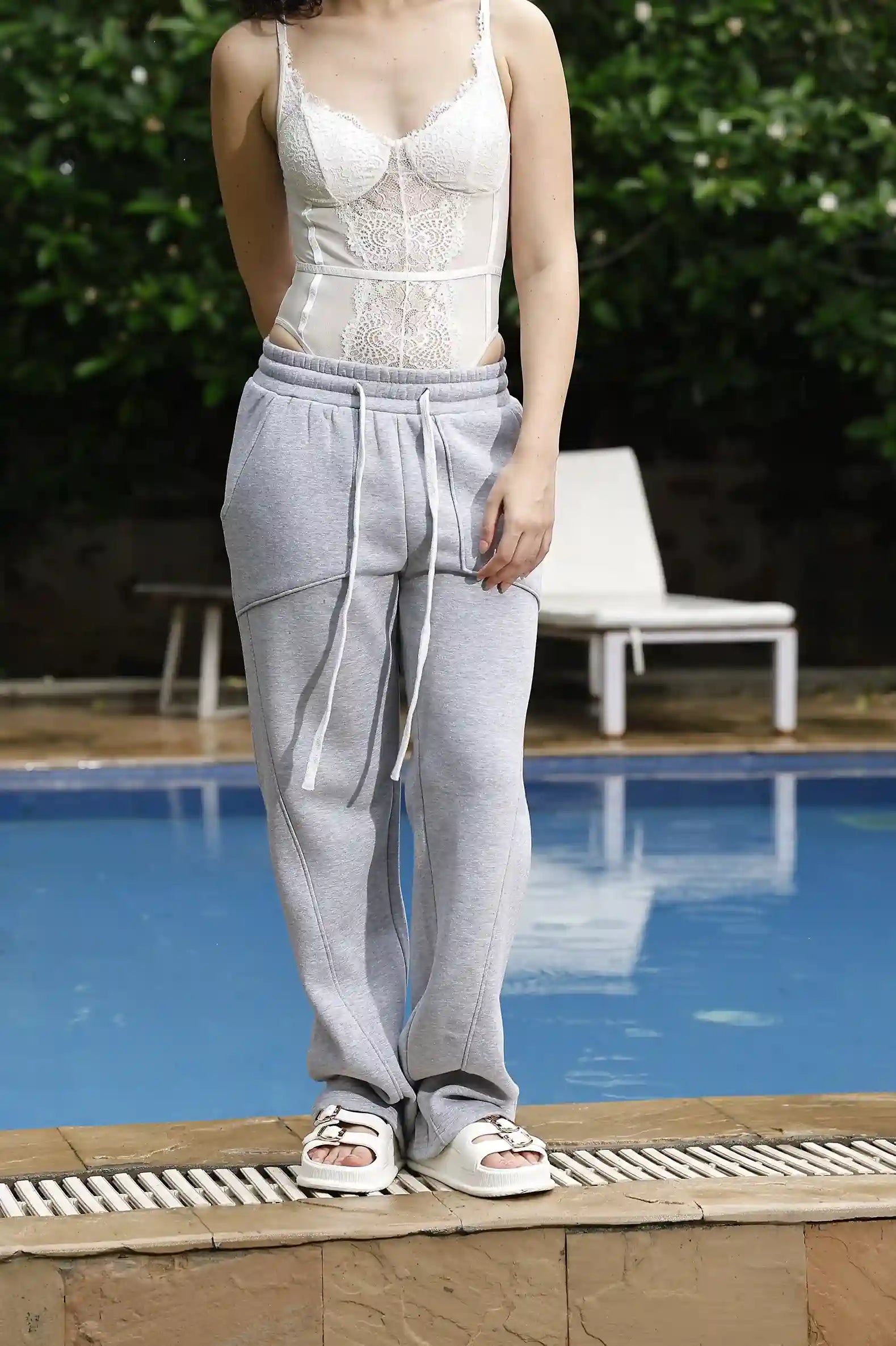 Person wearing a white lace top and gray sweatpants standing by a pool.