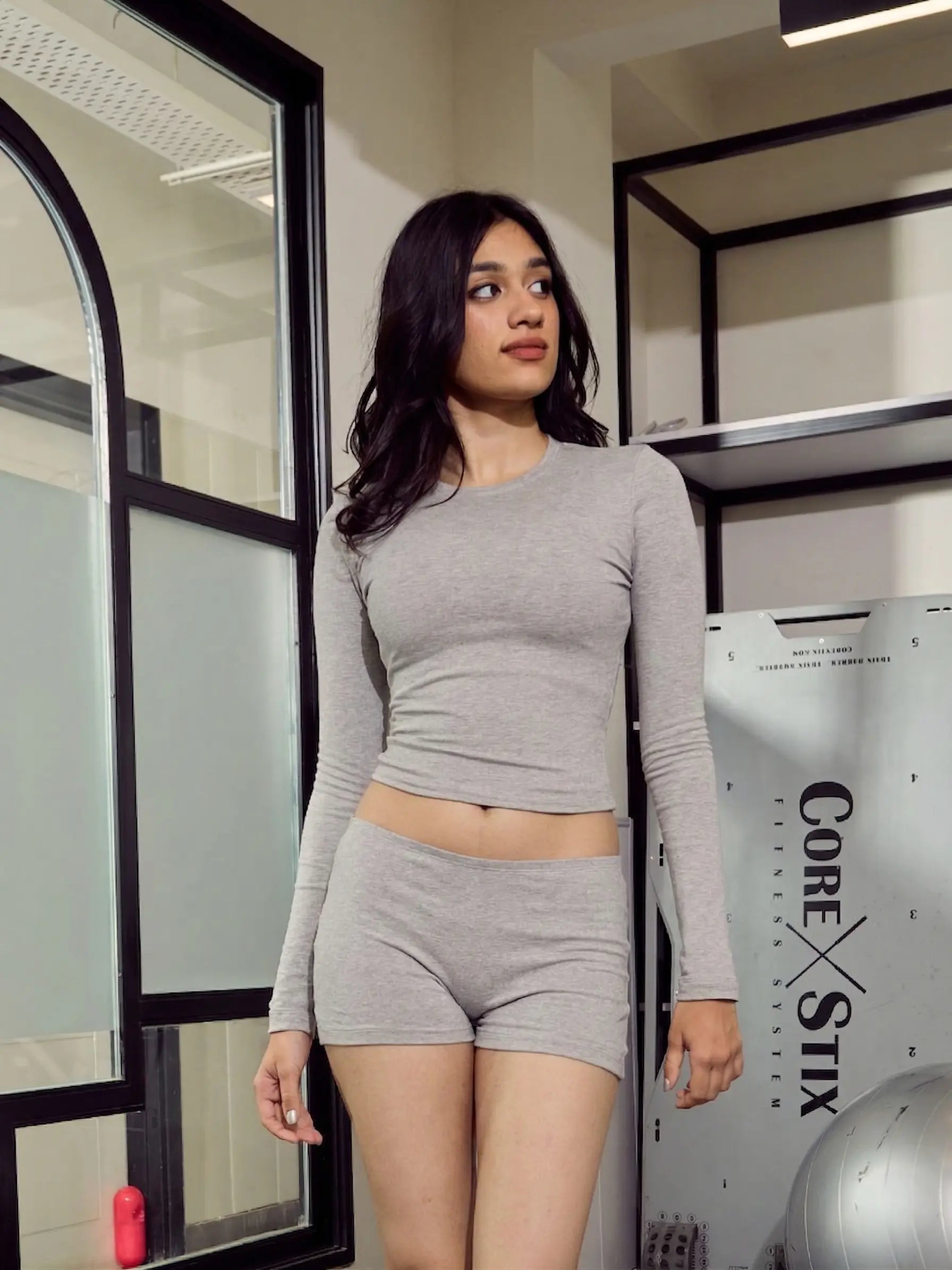 Woman wearing a grey long-sleeve top and shorts in a room with mirrors and a 'Core Stix' sign.