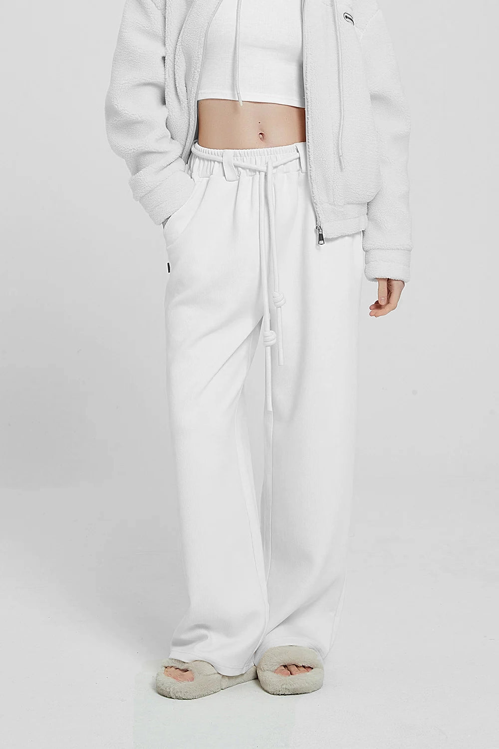 A person wearing white high waist knitted pants and a white hoodie with a logo on the sleeve, standing against a plain background.
