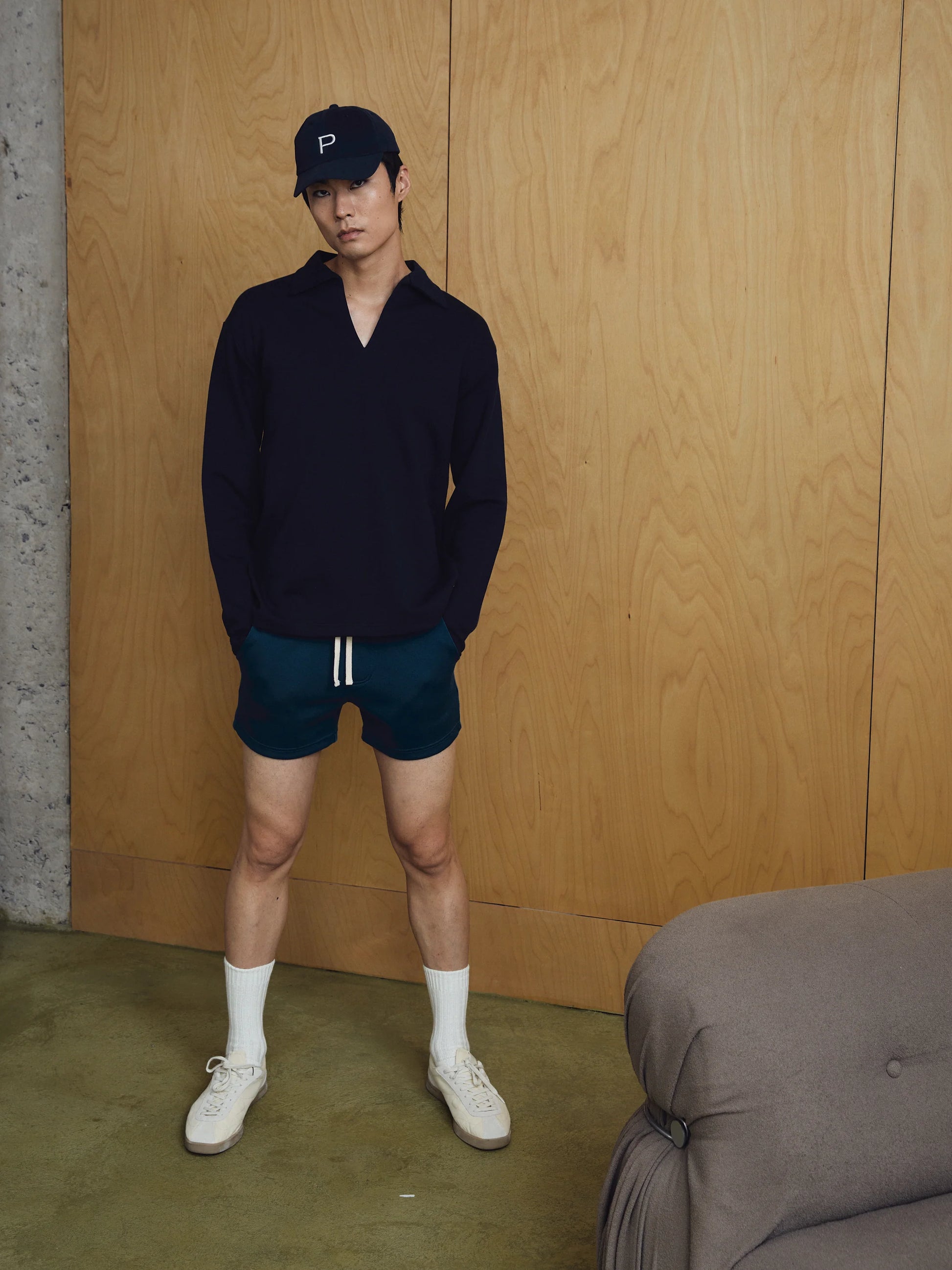 Person wearing a navy sweater, dark shorts, and white sneakers against a wooden wall.