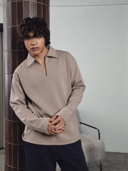 Person wearing a beige long-sleeve shirt standing in a room with a tiled wall and chair.