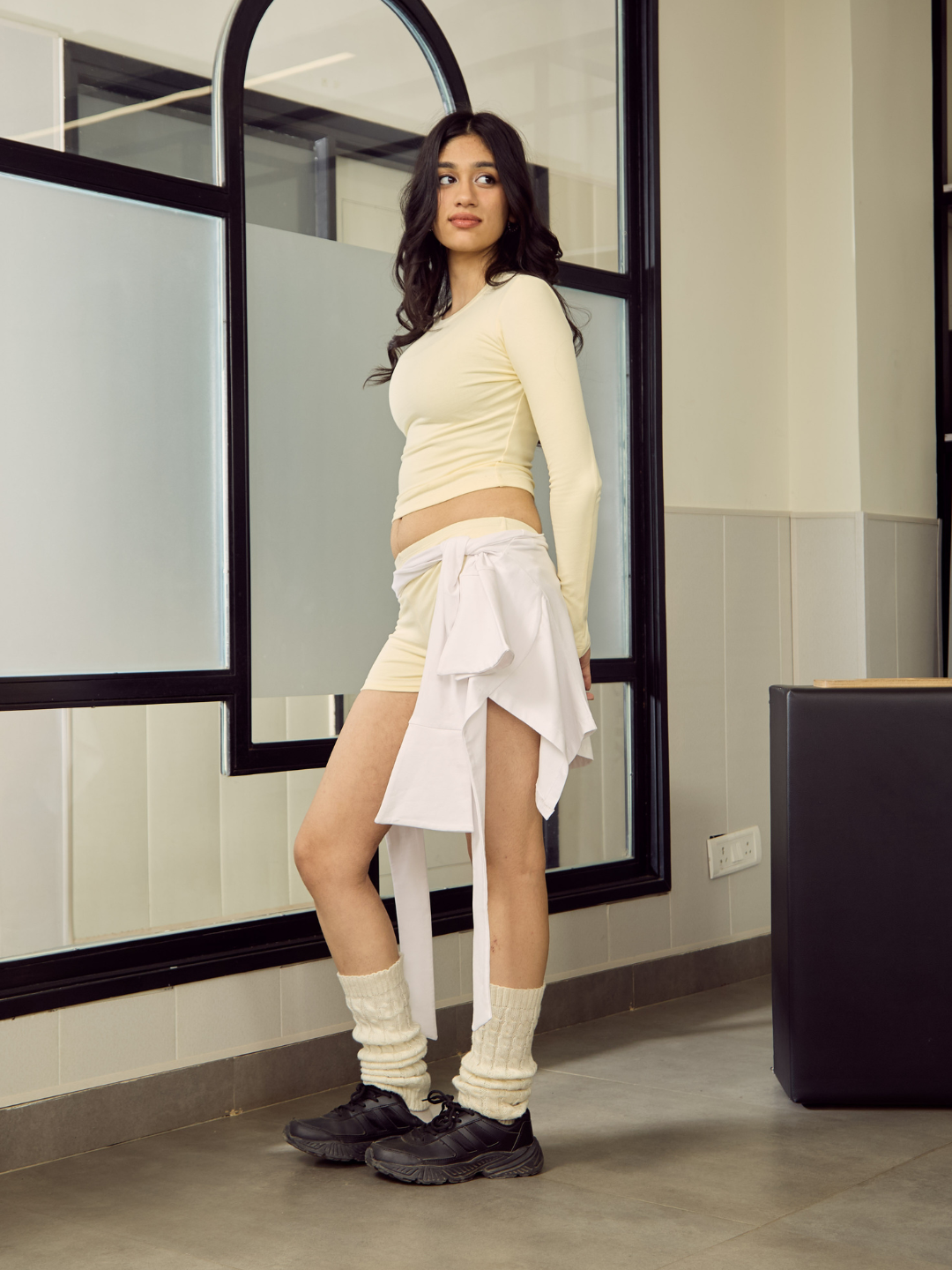 Woman in a yellow top and white skirt standing in front of a mirror.