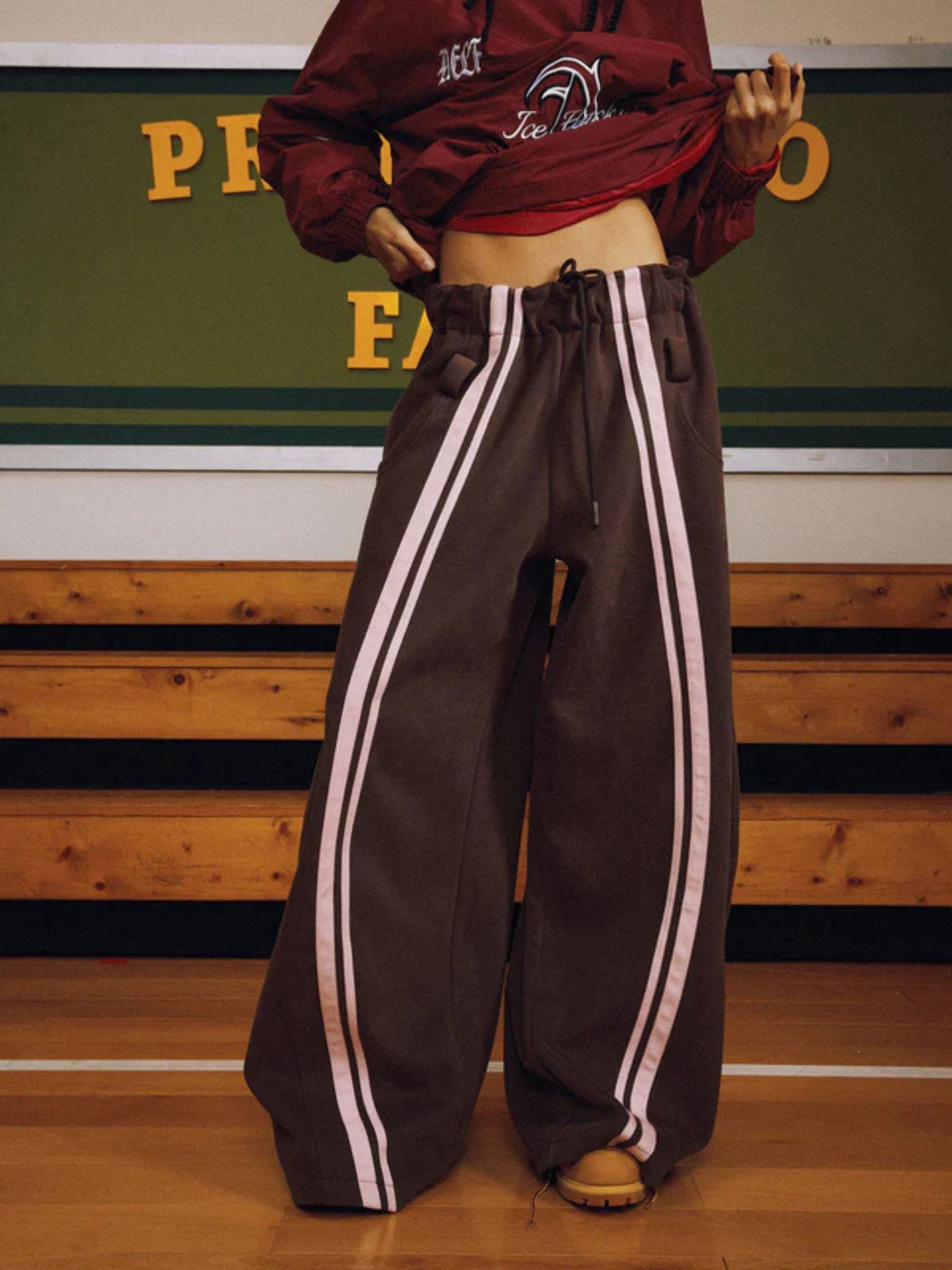 Person wearing a maroon jacket and brown track pants with white stripes on a gymnasium floor.