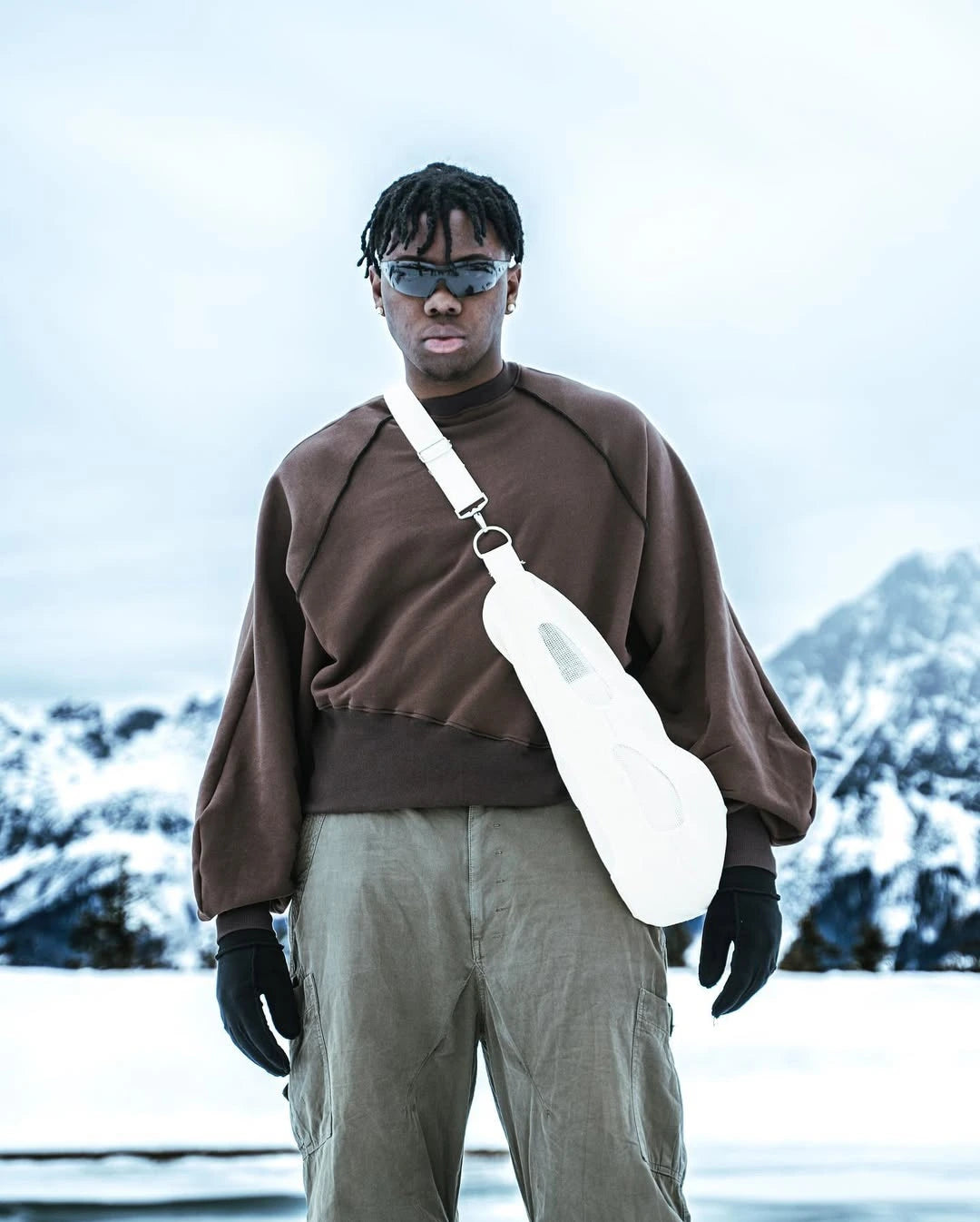 Person wearing a brown sweater and sunglasses, holding a white bag with a mountainous background