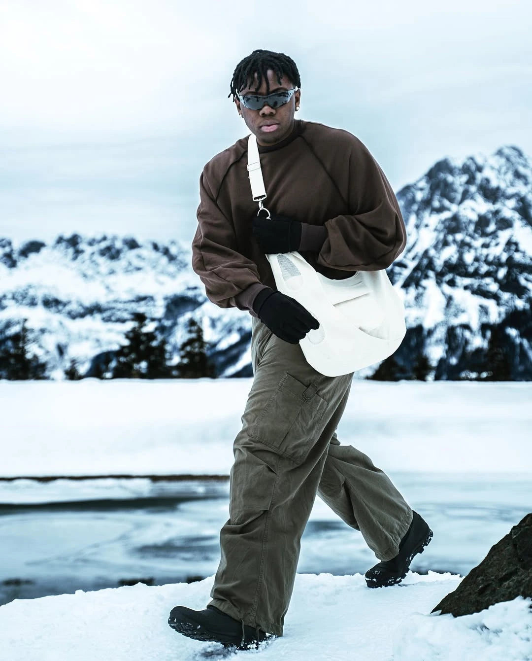 Person walking in a snowy landscape wearing a brown sweater and beige pants.