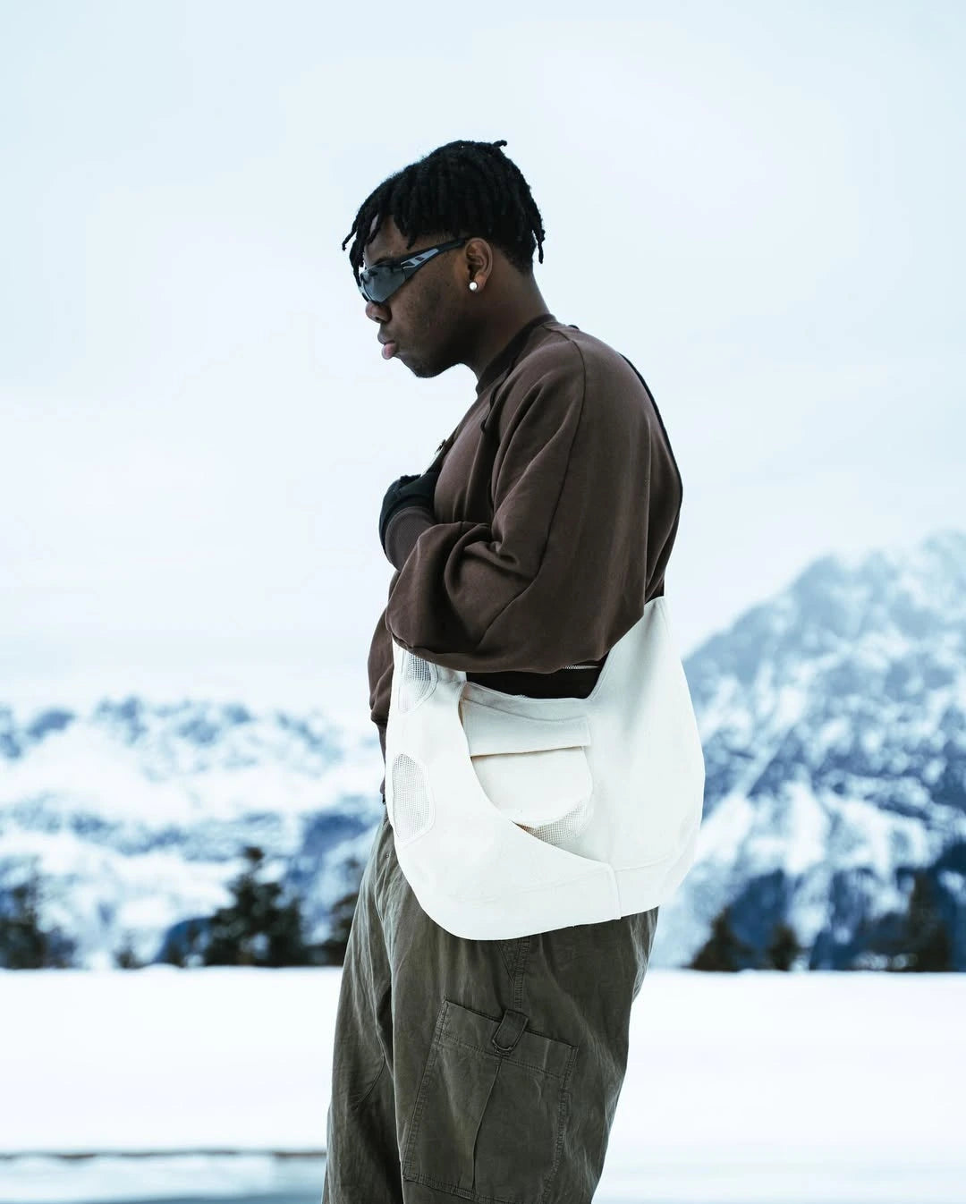Person wearing sunglasses and a brown jacket with a white bag in a snowy landscape