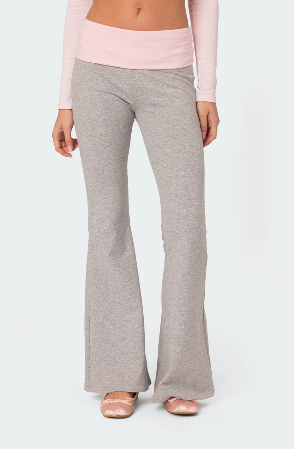 A female model wearing a pink rhinestone top with  long sleeves, paired with gray flare leggings. She is also holding a small white purse.