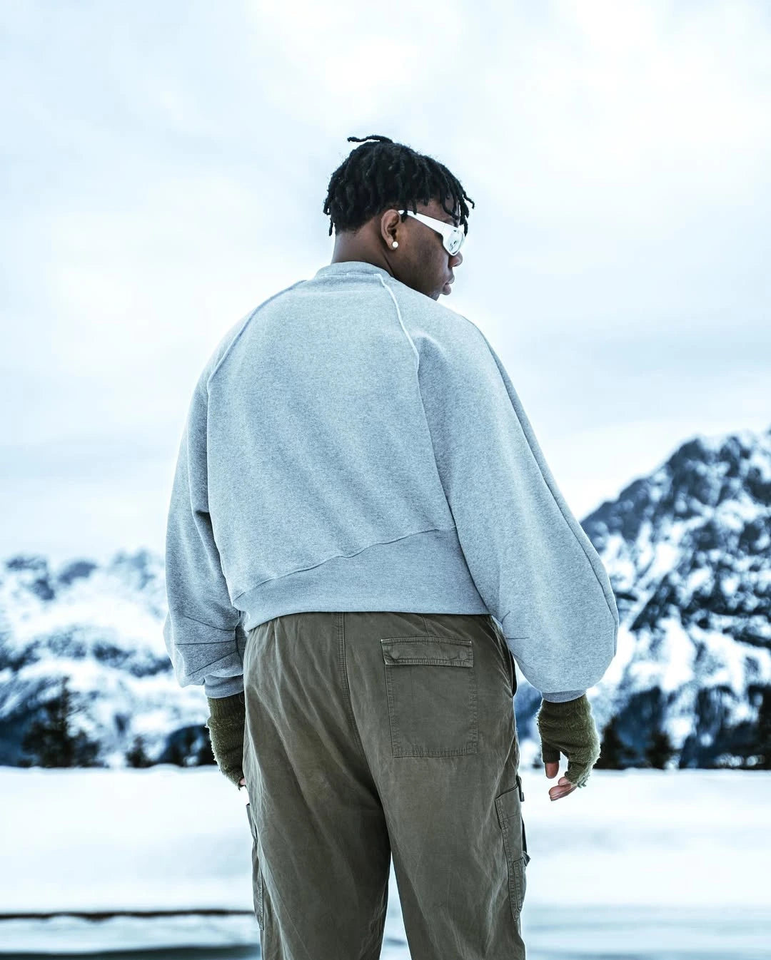 Person wearing a light gray sweatshirt and beige pants standing in a snowy landscape with mountains in the background.
