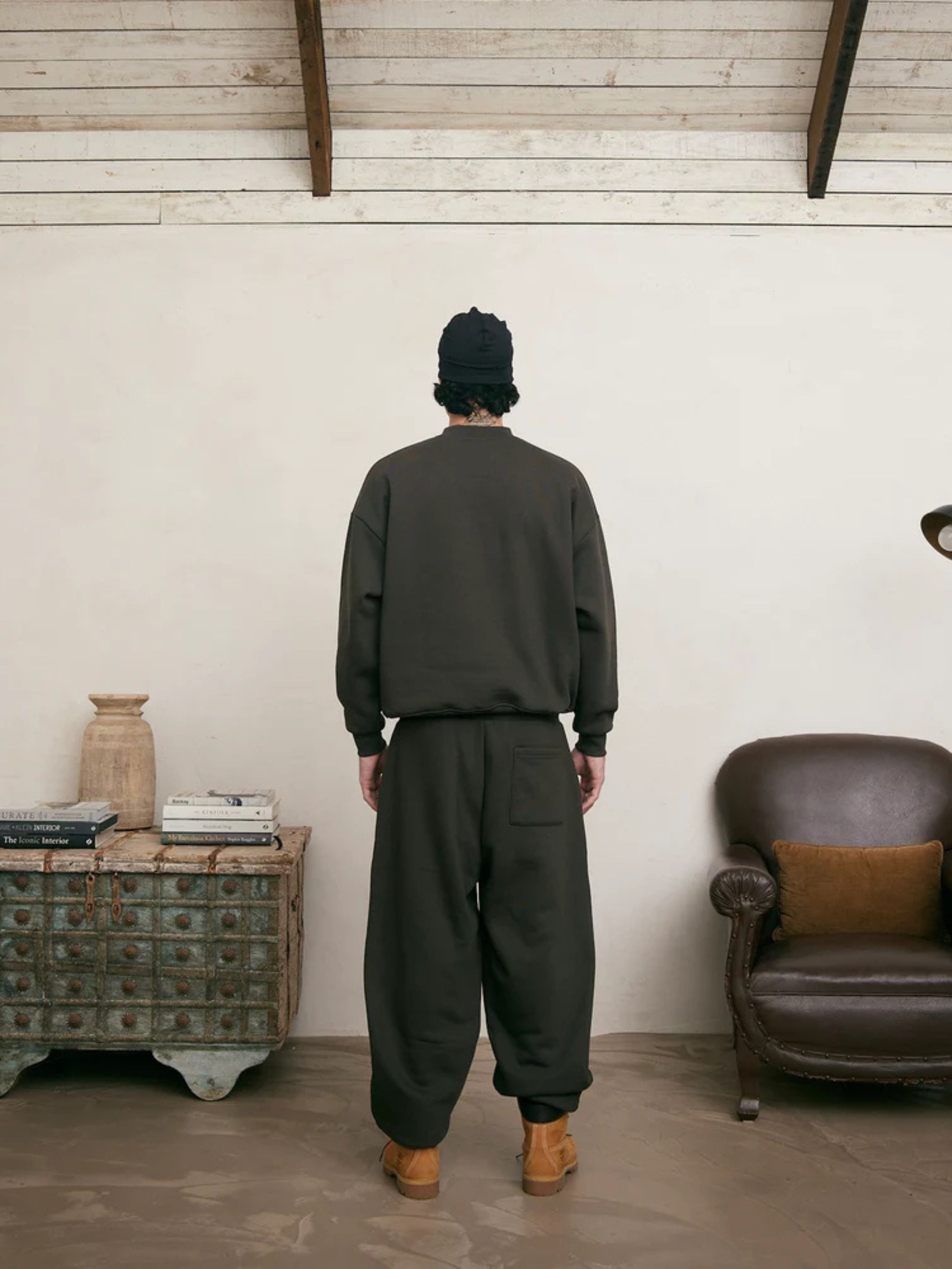 Person wearing a dark green outfit standing in a room with a leather chair and wooden cabinet.