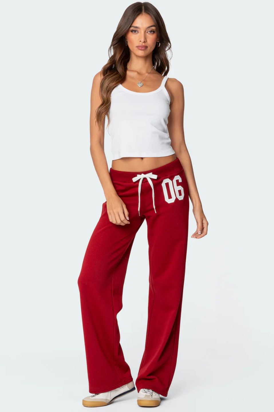 A female model wearing a white tank top and red lowrise trackpants with the number '06' printed on the left leg.