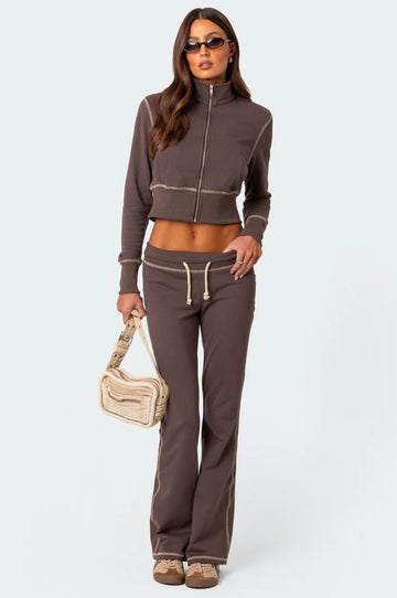 A woman standing and modeling a brown fleece club tracksuit, consisting of long-sleeved top and pants, with a white designer bag in her left hand.