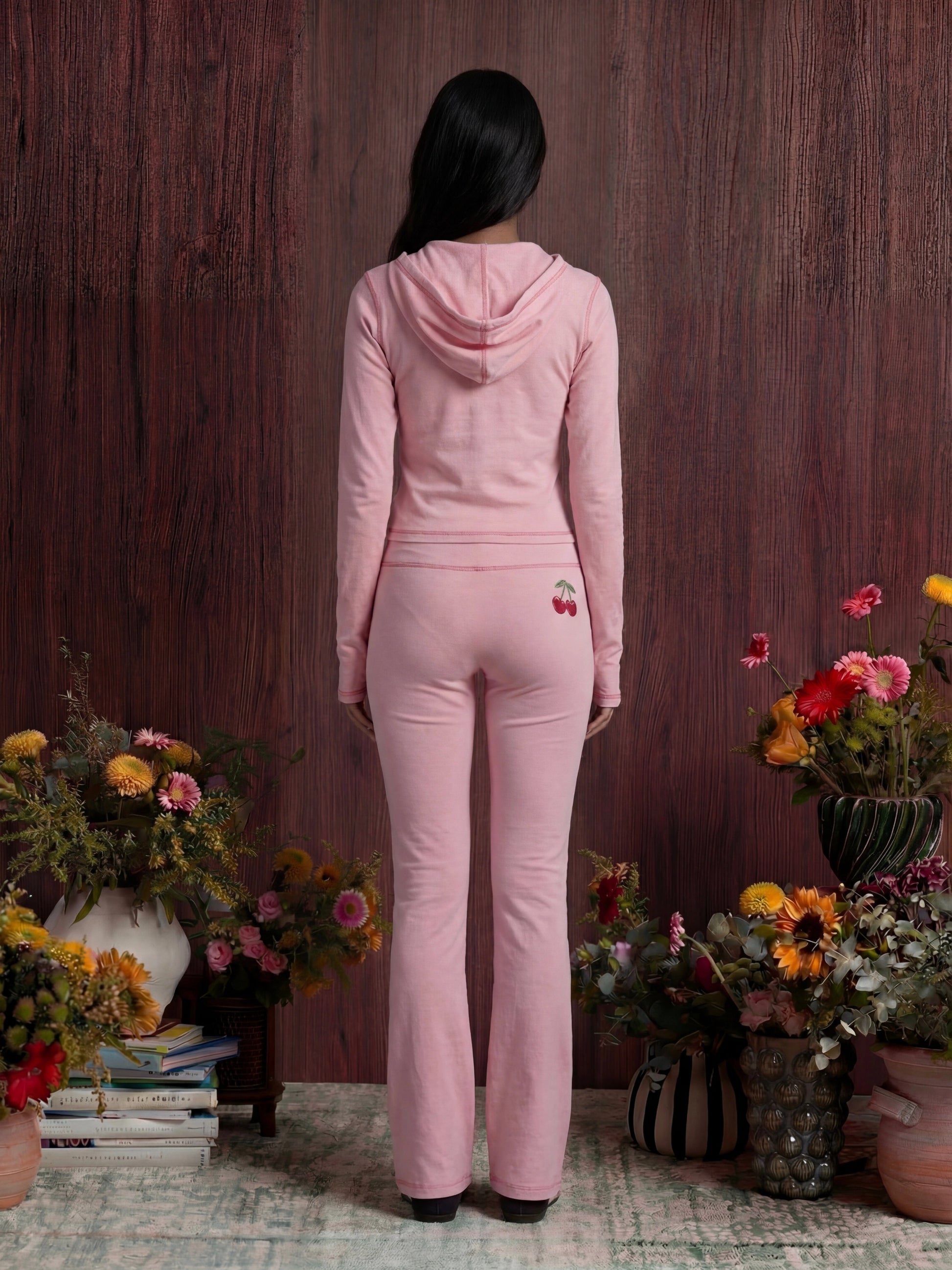 Woman in a pink outfit standing among floral arrangements against a wooden wall.