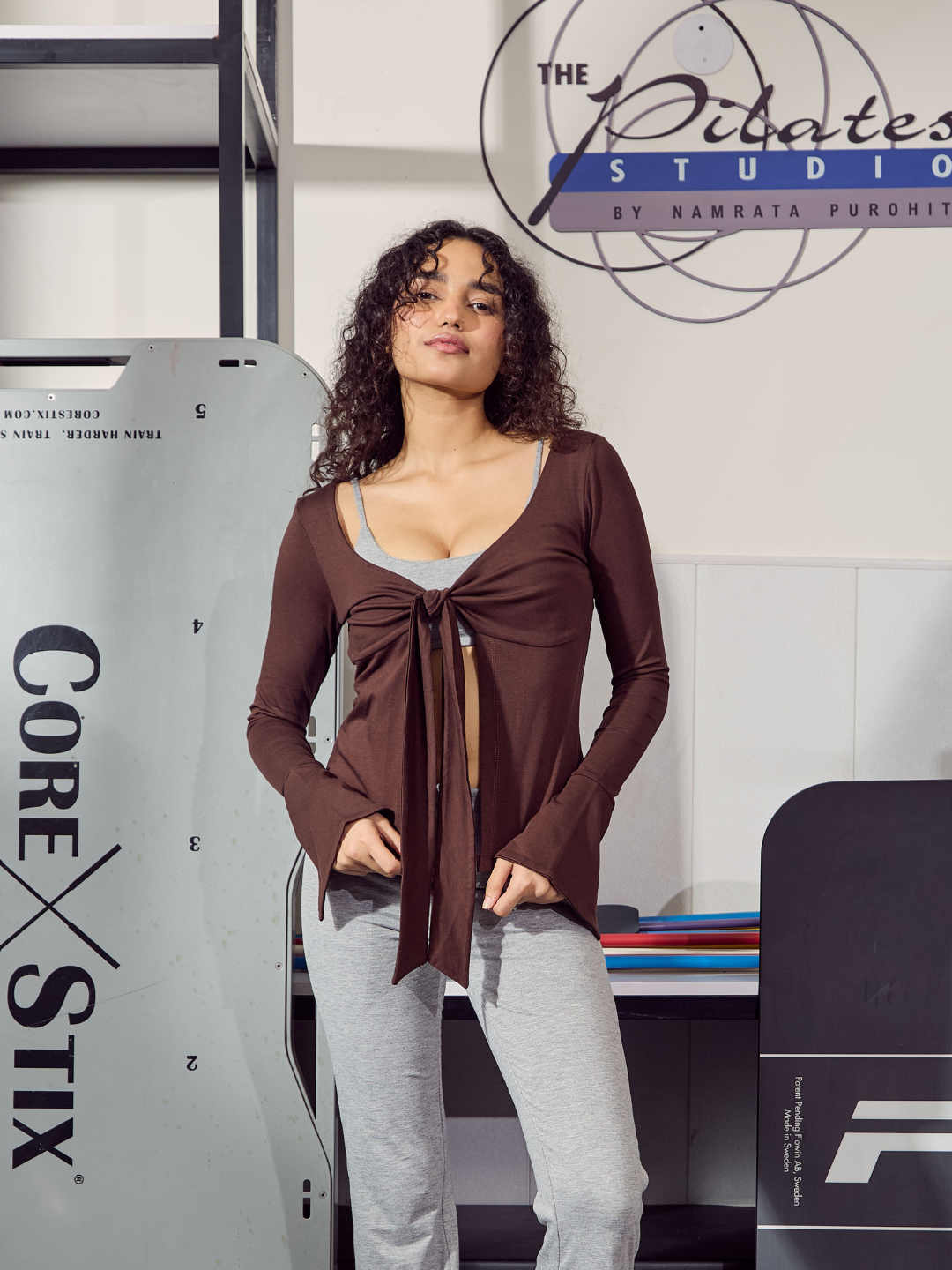 Woman standing in a Pilates studio with equipment and 'The Pilates Studio' branding.