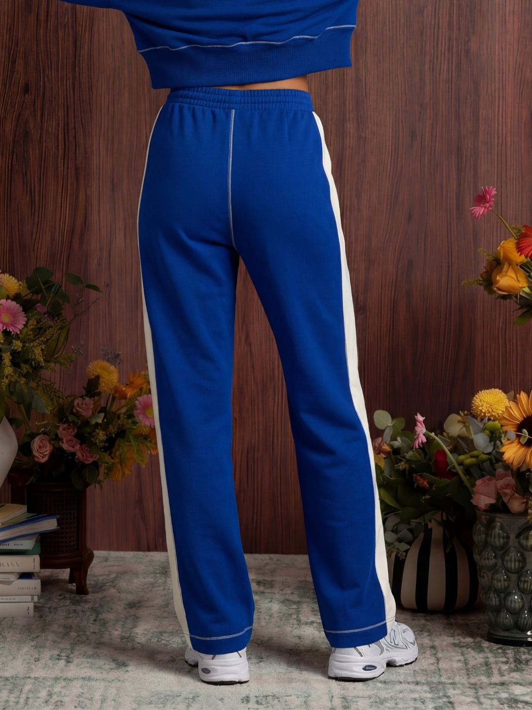 Person wearing a blue tracksuit with white stripes in a room with flowers and books.