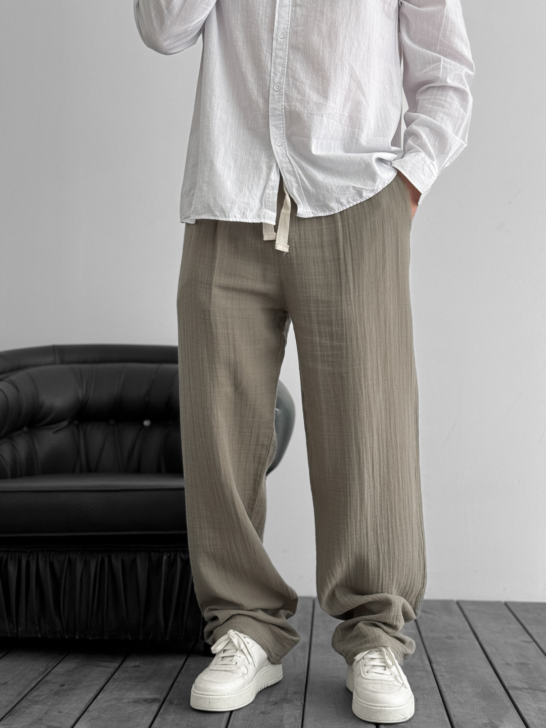 A person standing against a white background, wearing wide-leg gray gauze pants and a white shirt with the sleeves rolled up to the elbows.