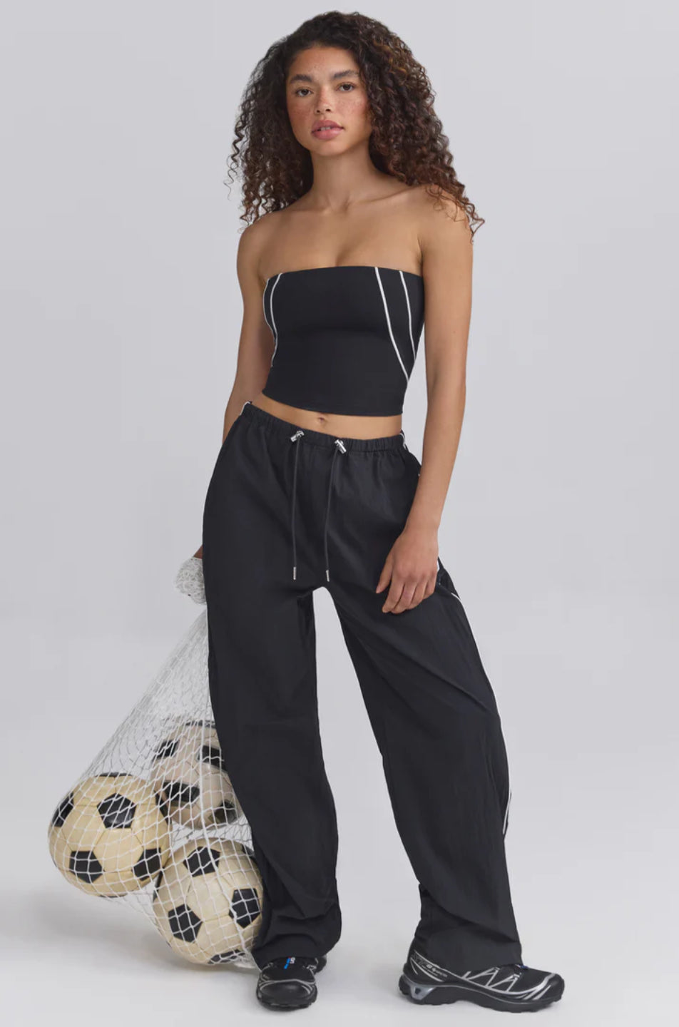 A woman standing and holding a soccer ball, wearing black wide-leg track pants with an elasticated drawstring waistband and contrast piping.