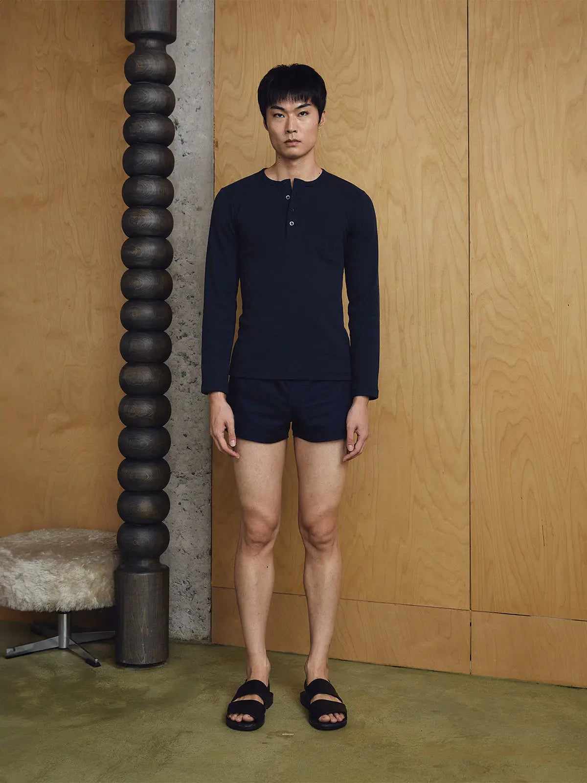 Man wearing a navy long-sleeve shirt and shorts standing against a wooden wall.